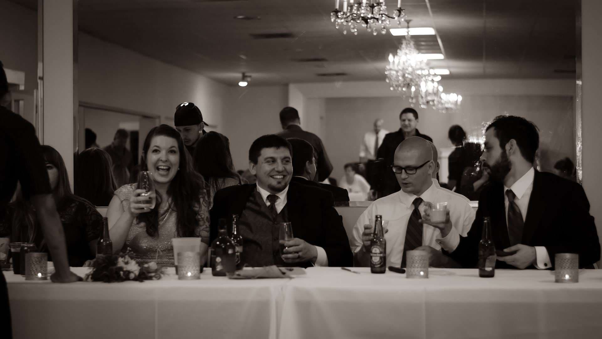 Sepia image of wedding guests laughing and holding drinks at a long white-clothed reception table under chandeliers