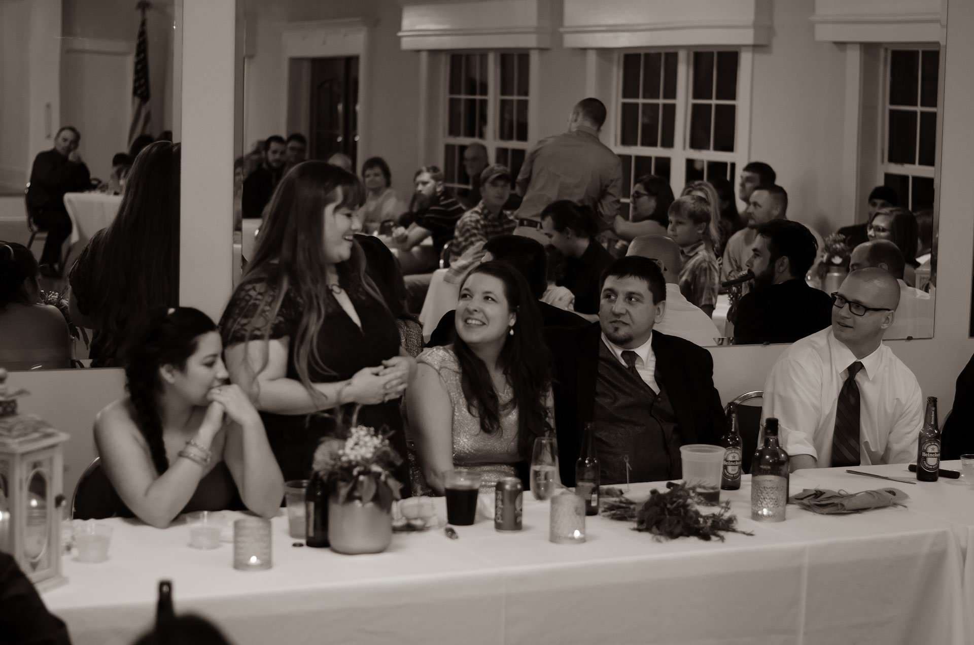 Sepia photo of wedding guests seated at a long reception table, a standing woman talking while others smile up at her