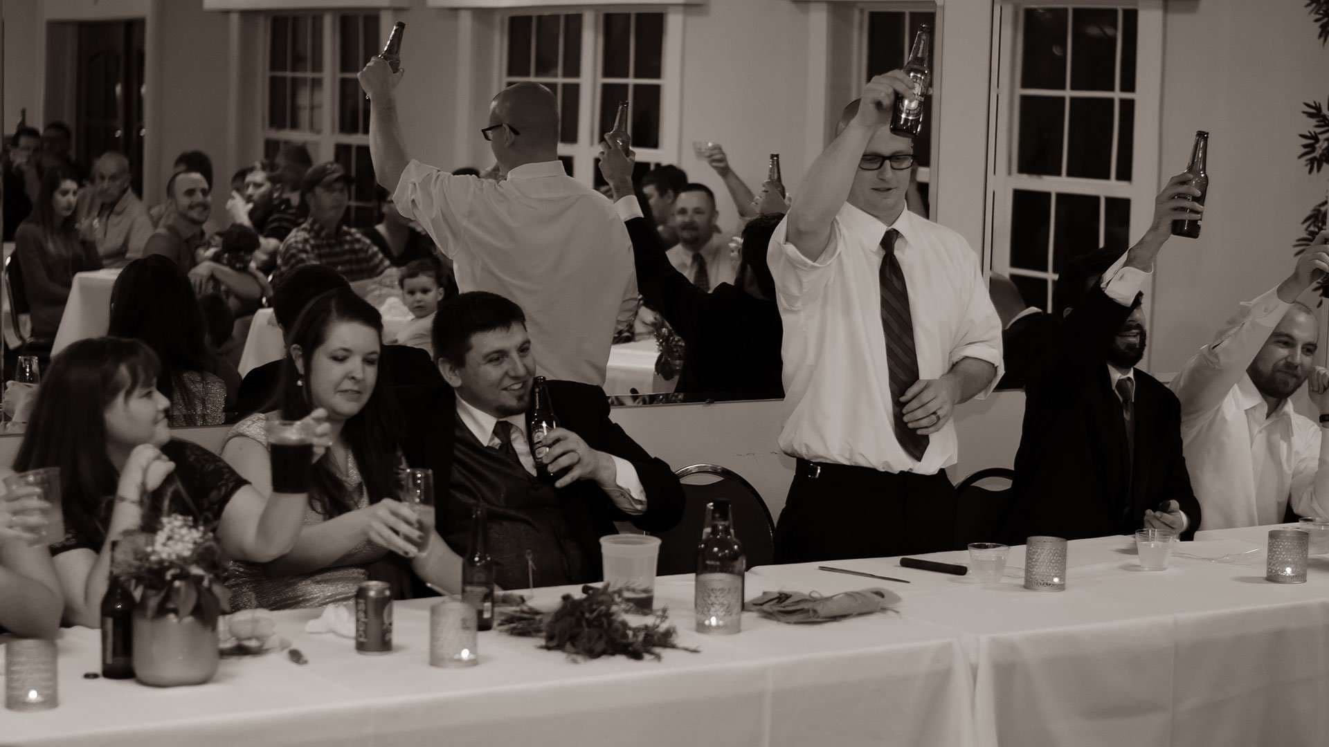 Sepia photo of wedding guests standing and raising beer bottles in a toast at a reception table