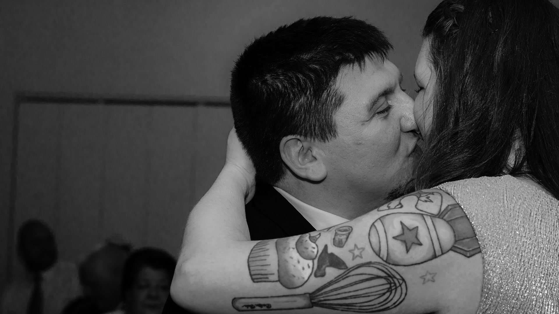 Black-and-white close-up of a couple kissing, her forearm tattooed with a whisk, cupcake and star designs