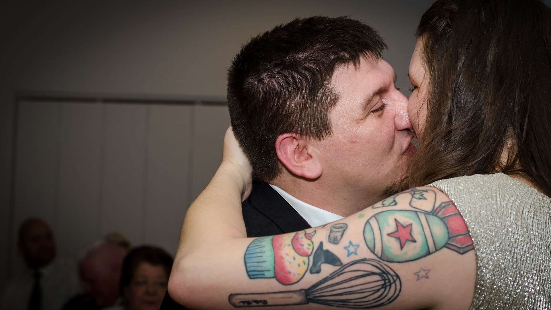Couple kissing during an embrace, her forearm showing colorful tattoos of a whisk, cupcake and star-marked bomb