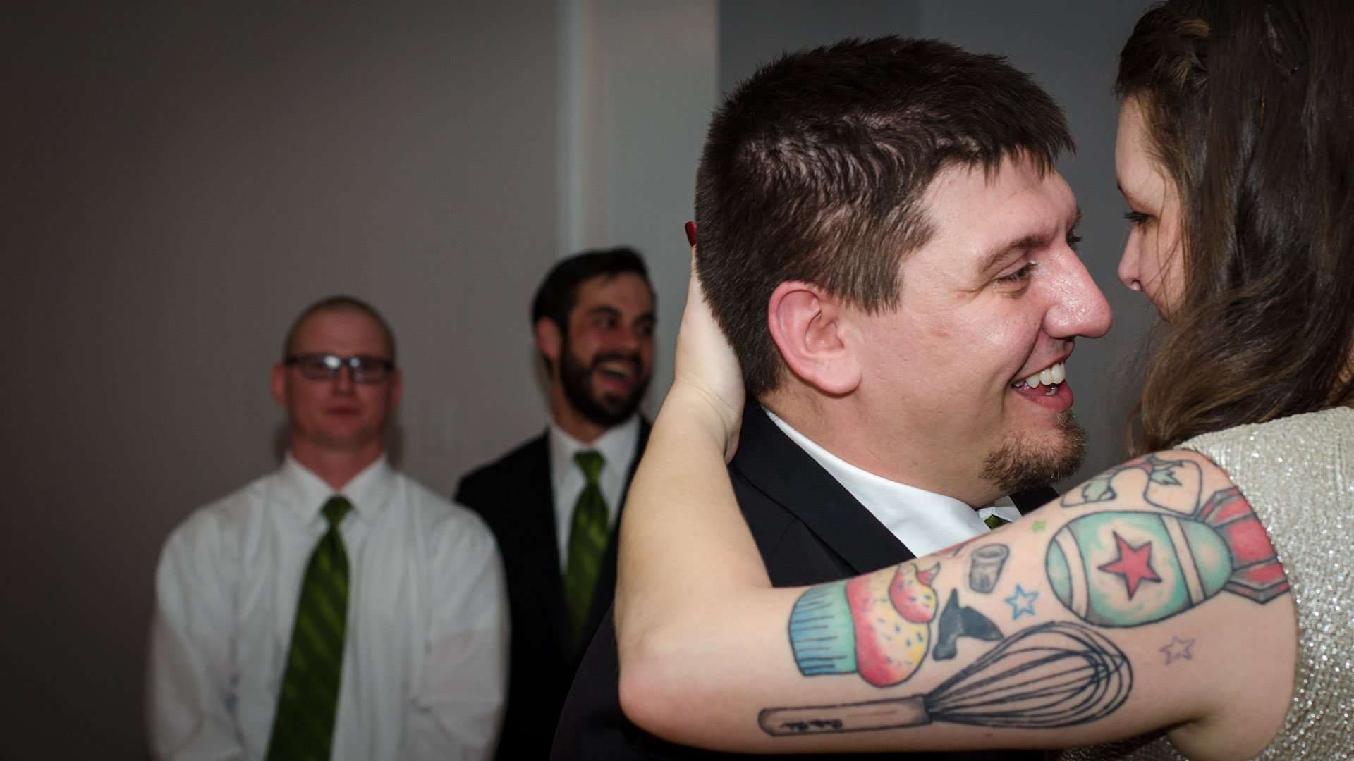 Smiling groom in a dark suit embraced by a tattooed woman while two groomsmen in green ties laugh behind them