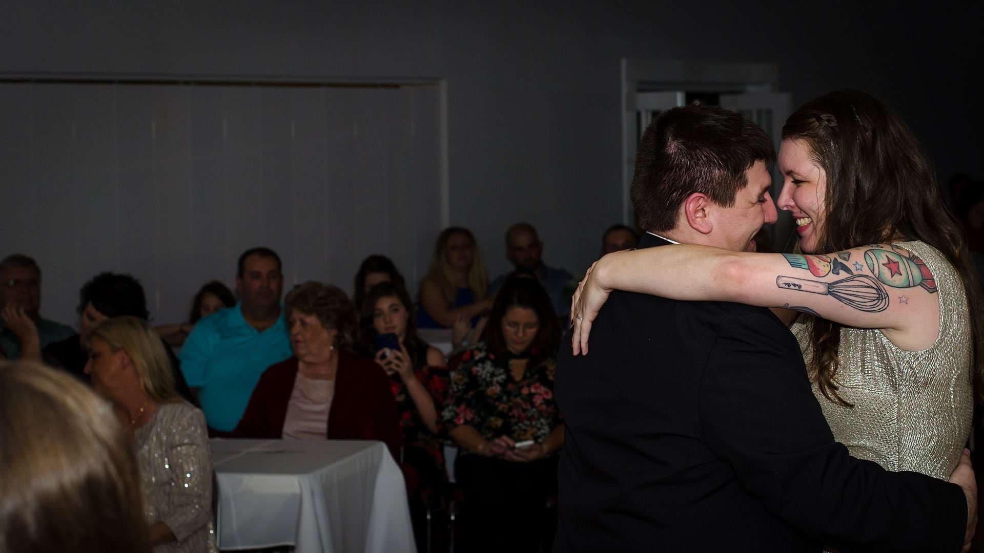 Smiling woman with tattooed arm embraces a man in a dark suit while seated guests watch behind them