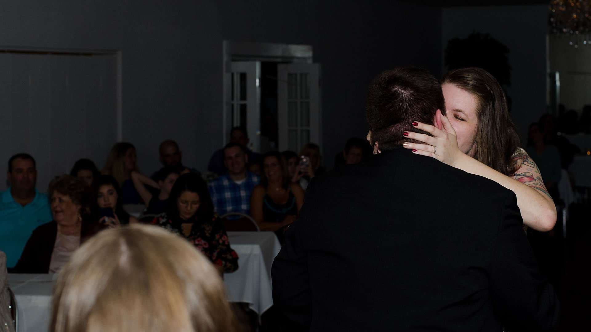 Couple embracing and kissing in a dim reception hall while seated guests watch from tables behind them