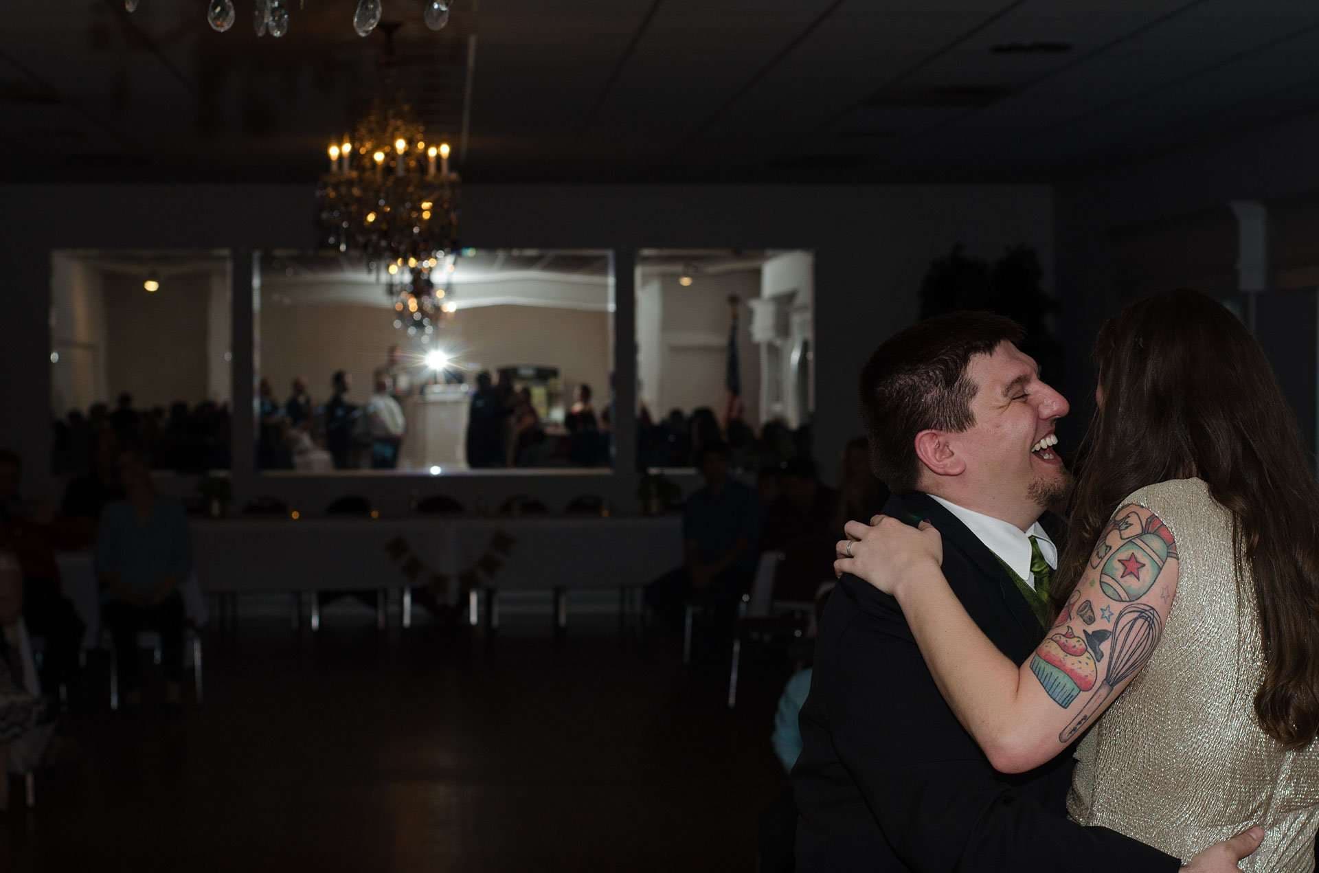 Groom laughing as he dances with his tattooed bride under a chandelier at the reception