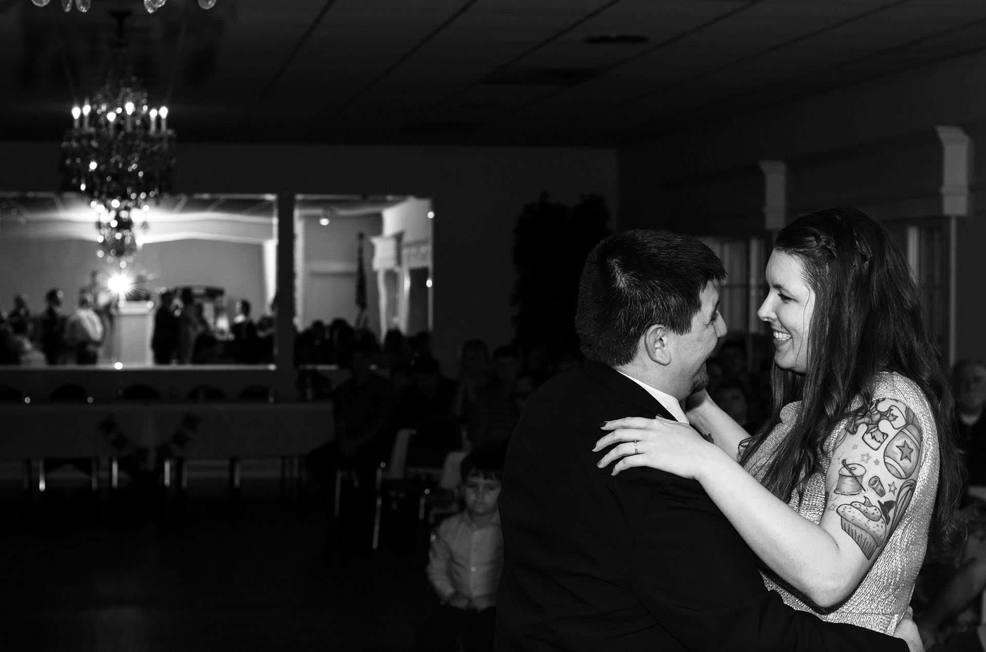 Black-and-white shot of a smiling tattooed bride dancing with her groom in a chandelier-lit hall as guests watch