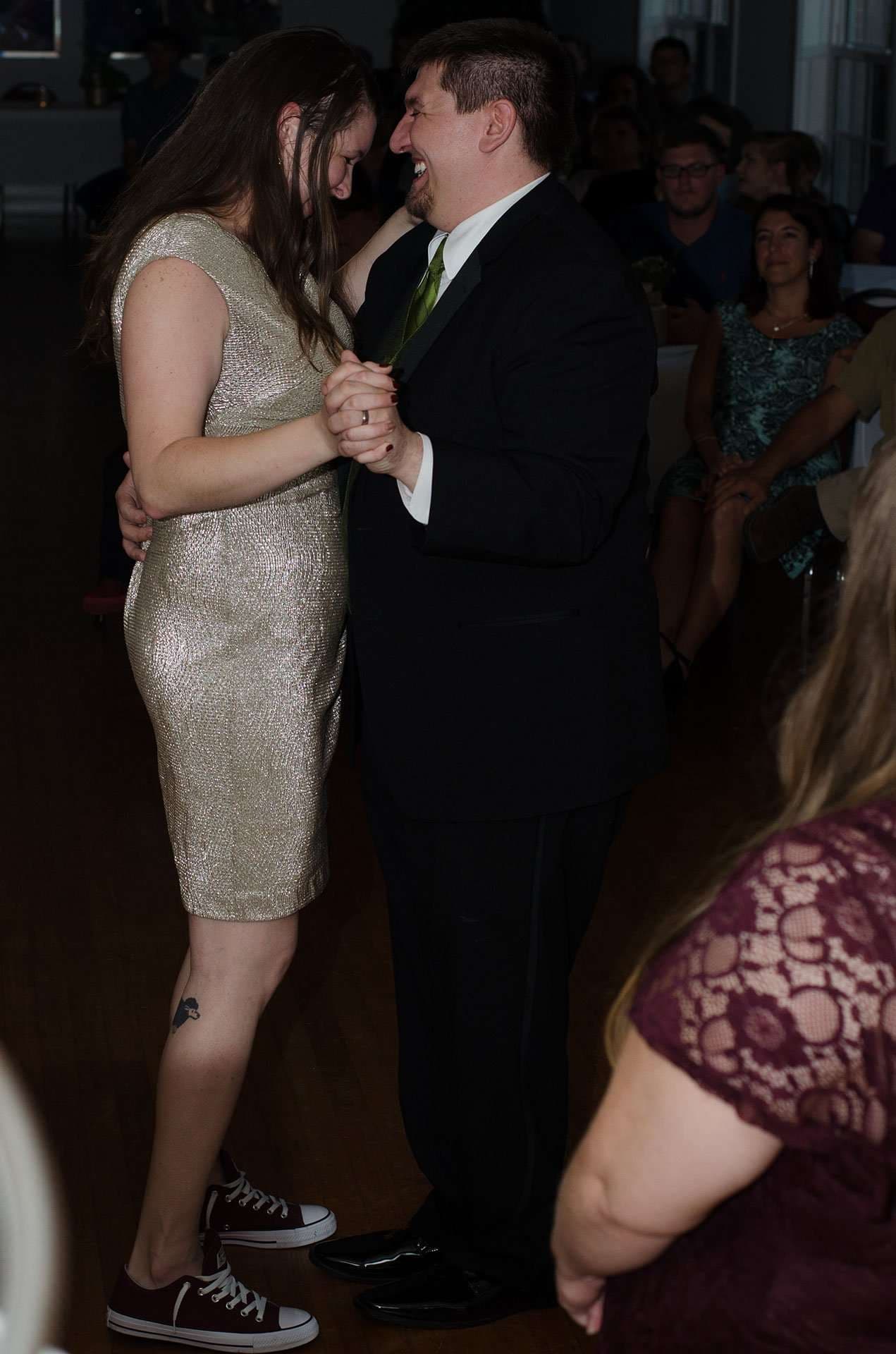 Couple laughing and slow dancing on a wood floor, she in a gold sequin dress and sneakers, he in a dark suit