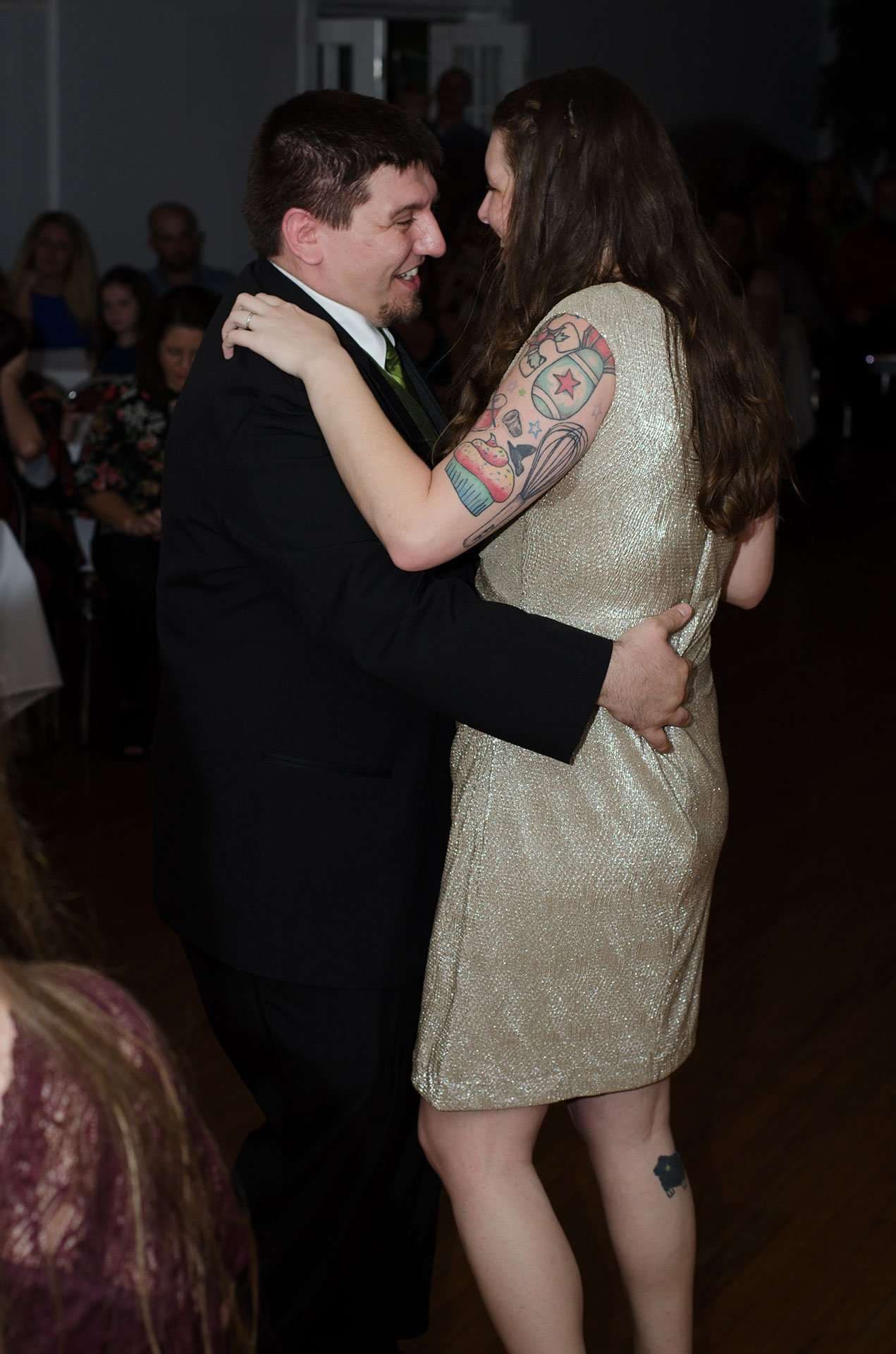 Couple slow dancing at a wedding reception, woman in a gold sequin dress with a colorful arm tattoo, man in dark suit