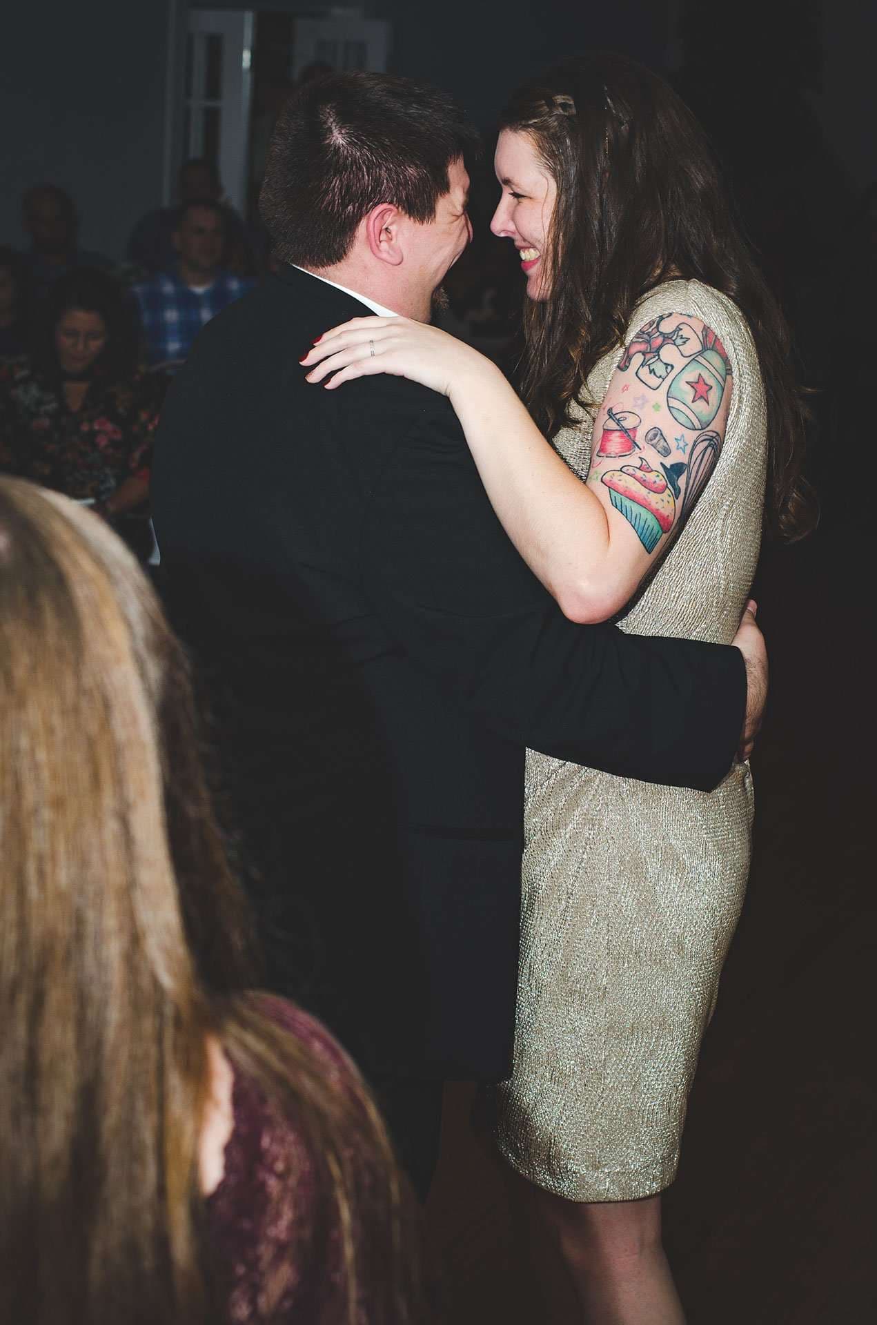 Smiling couple embracing during a dance, the woman in a gold dress with a colorful sleeve tattoo, guests watching behind