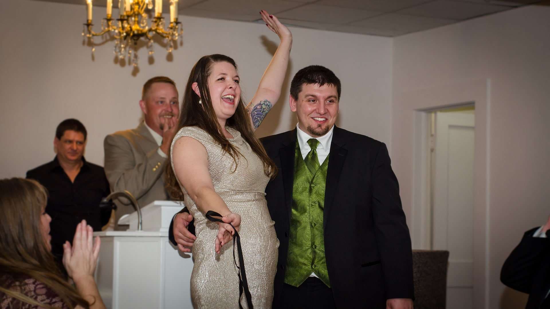 Laughing woman in a gold dress raises her arm beside a smiling man in a green vest as guests clap