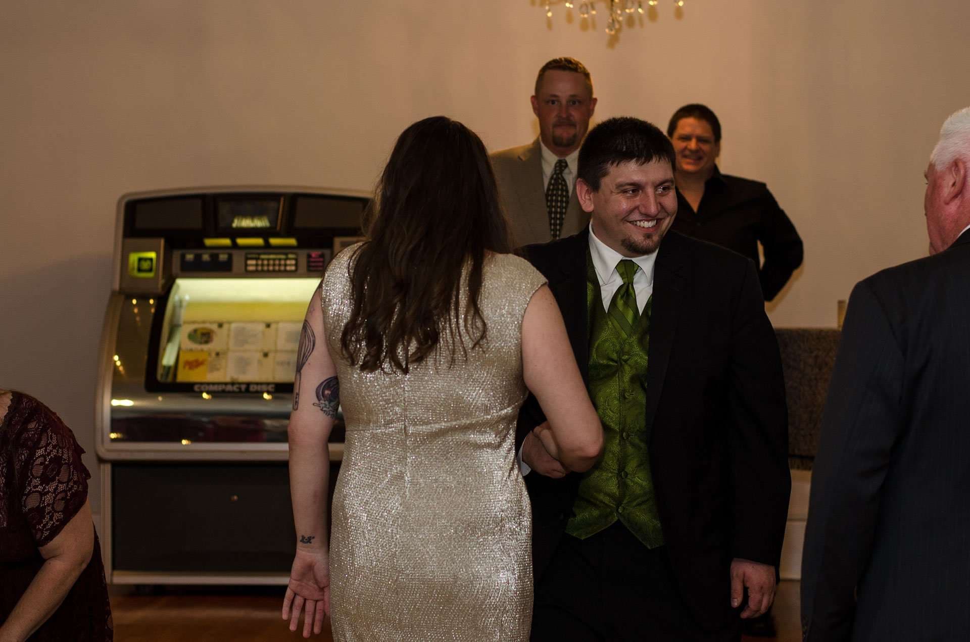 Smiling man in a green vest and tie greets a woman in a sparkly gold dress near a vintage jukebox