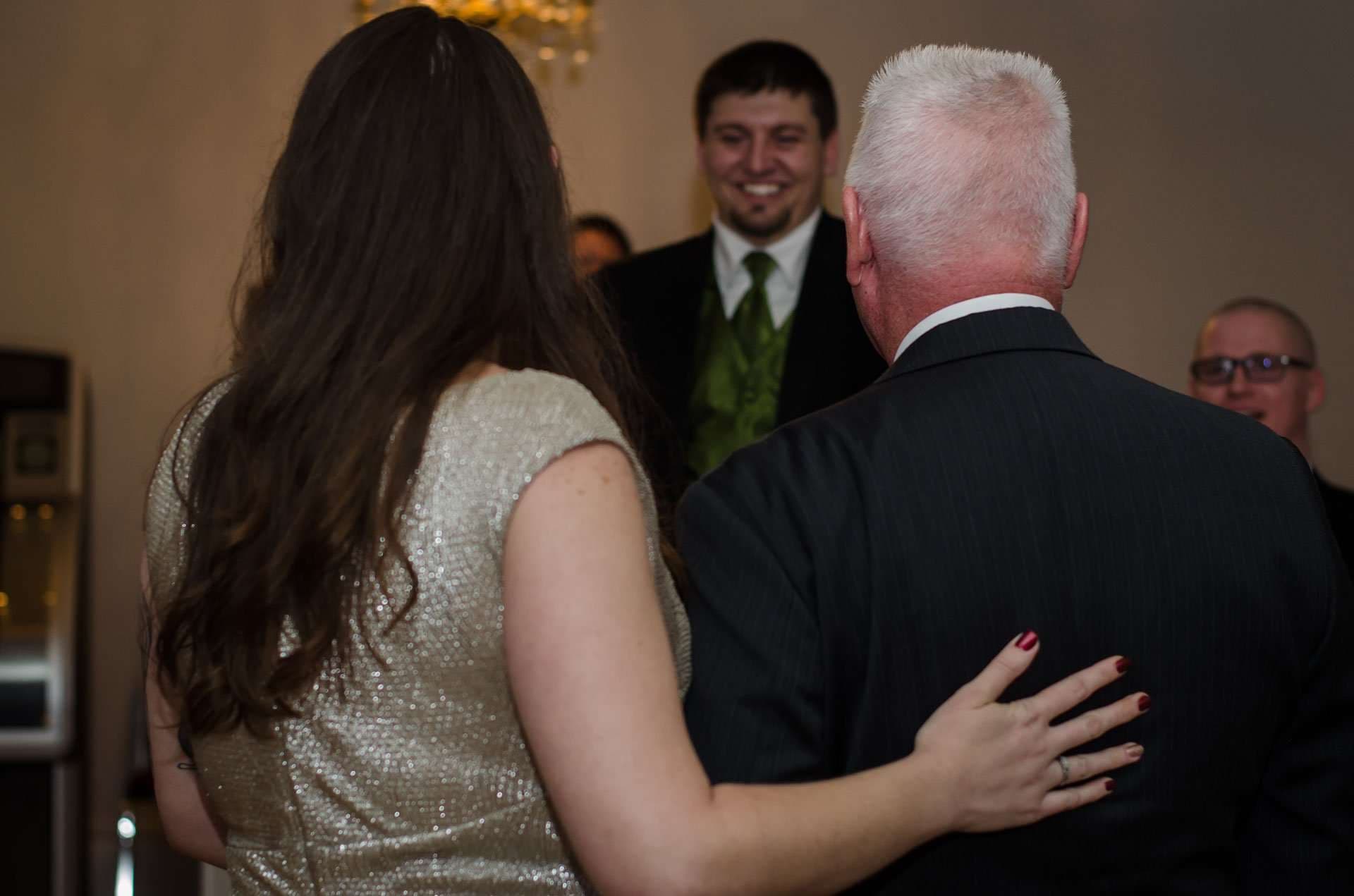 Woman in a sparkly gold dress rests her hand on an older man's back as a smiling groom in a green vest looks on