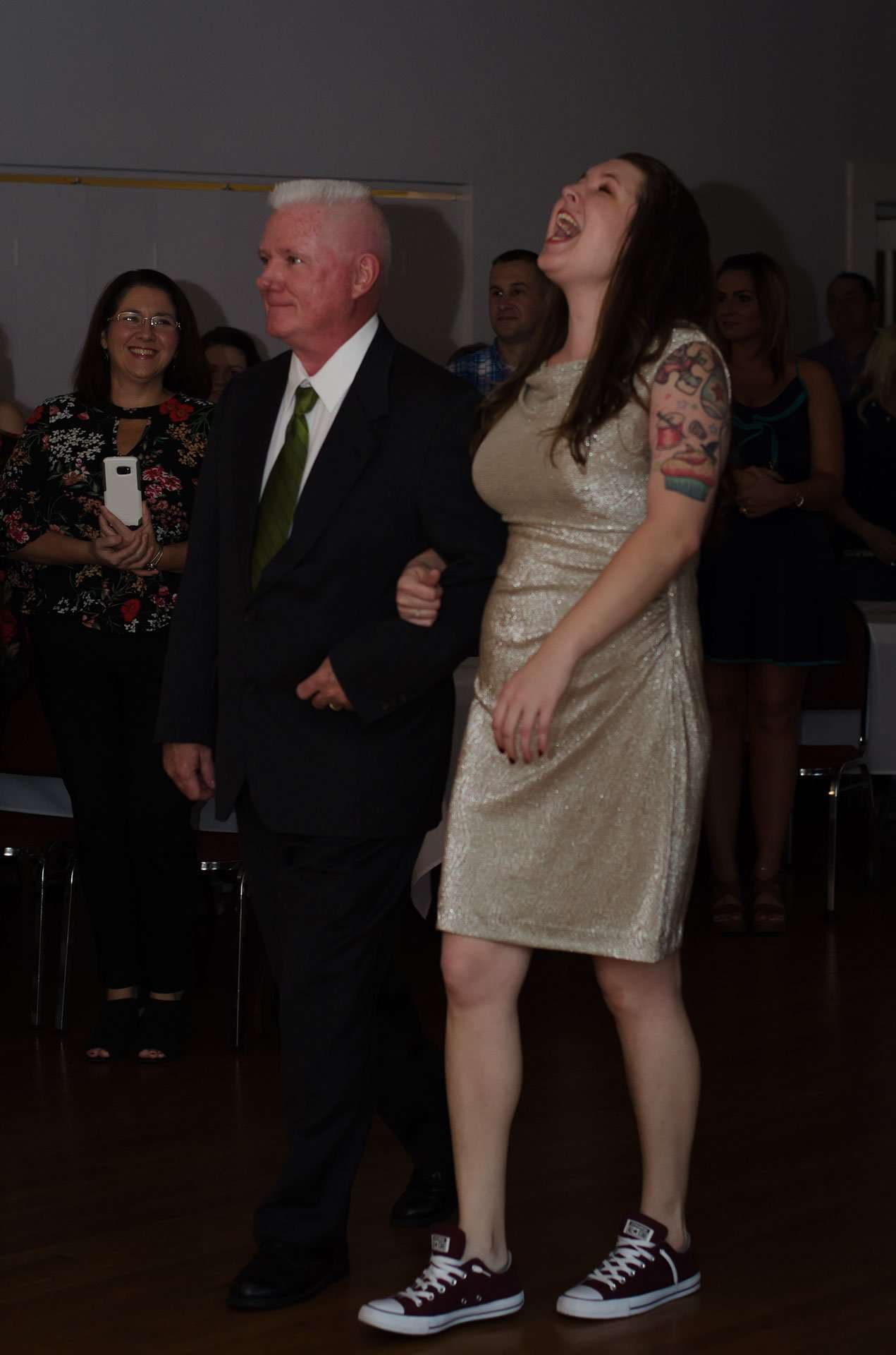 Older man in a dark suit and green tie walks arm in arm with a laughing woman in a gold dress as guests watch