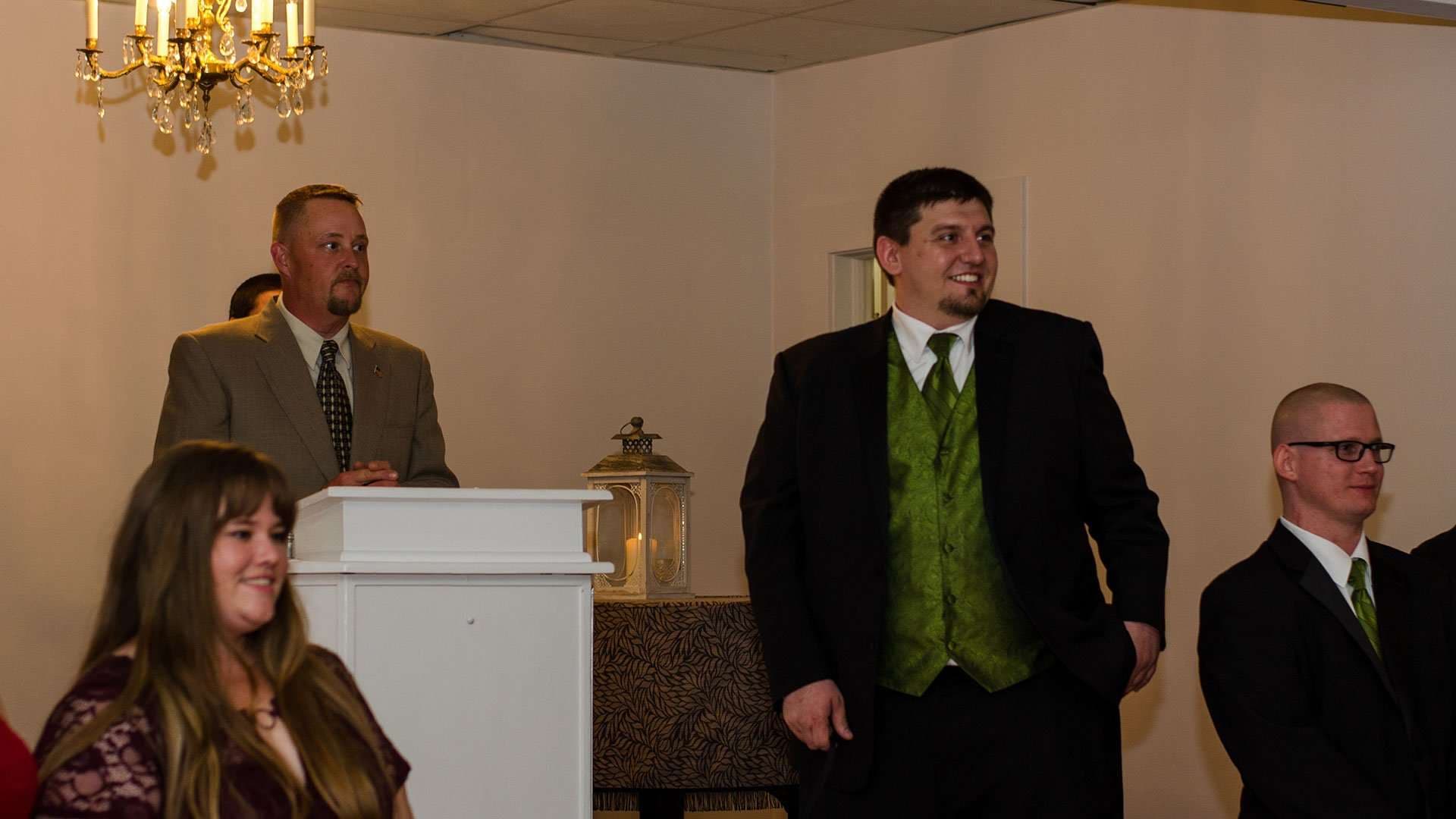 Groom in a green vest waiting at the front of the ceremony room beside the officiant