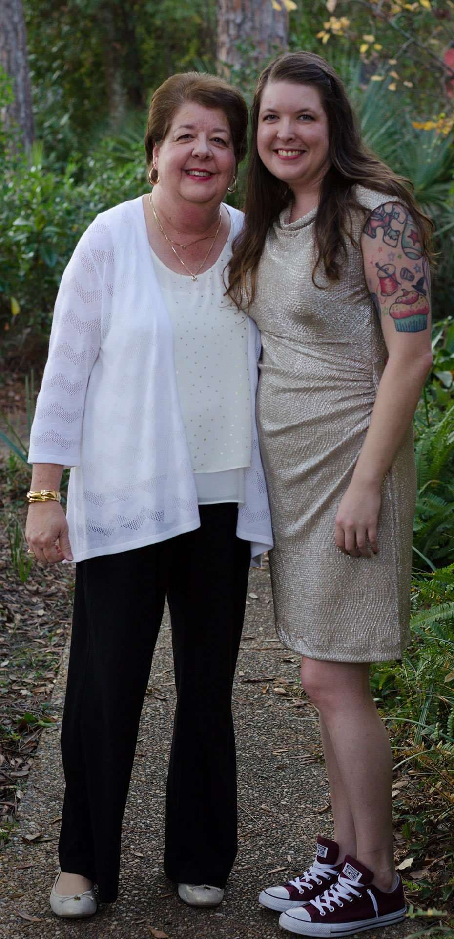 Older woman in white cardigan and younger woman in a gold dress with arm tattoos smiling together on a garden path