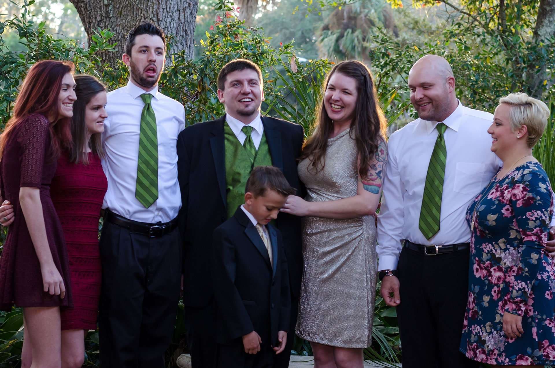 Smiling wedding party in green and black formal wear, a young boy in a suit at the center, outdoors in a garden