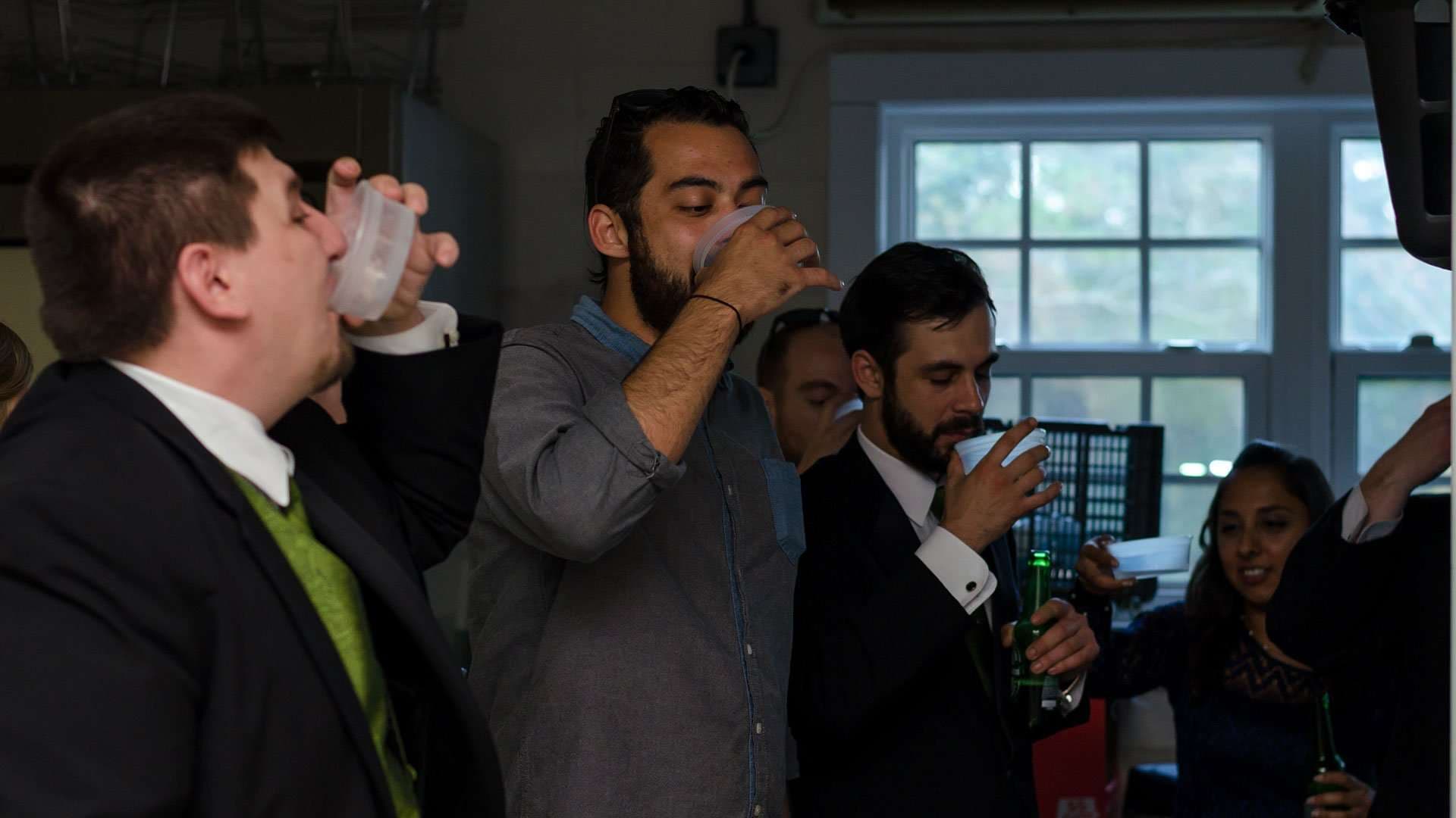 Men in suits and a gray shirt drinking from plastic cups at an indoor wedding gathering at the Panama City Women's Club