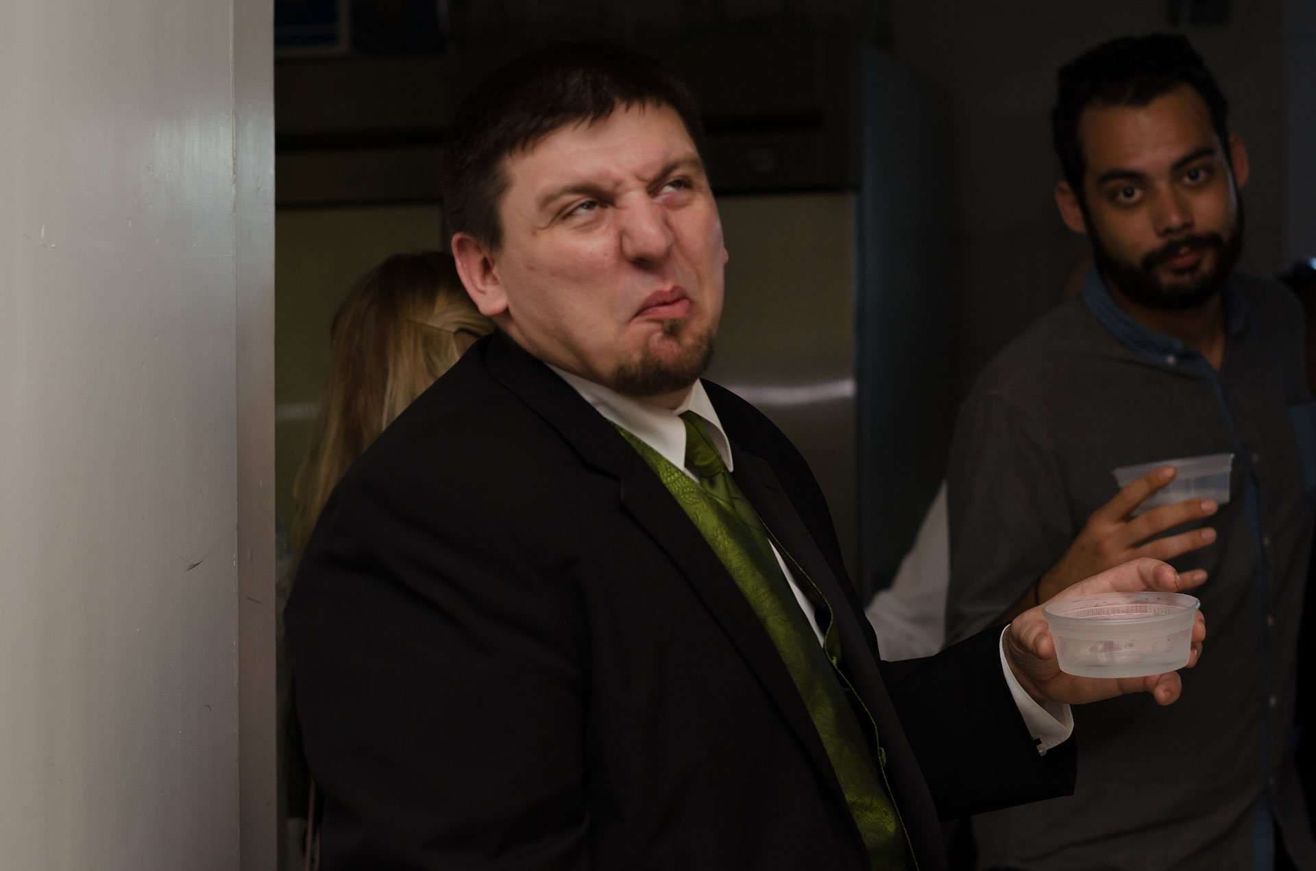 Man in a black suit and green vest scrunches his face humorously, holding a clear cup, as a bearded man looks on