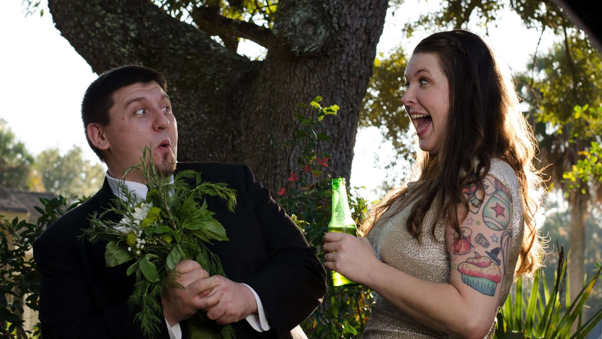 Groom holding a bouquet of greenery while his laughing bride holds a green bottle under an oak tree