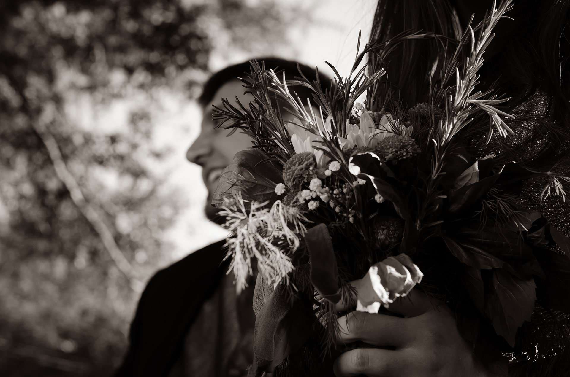 Sepia close-up of a rosemary and wildflower bouquet held in the foreground while a smiling man looks away, blurred behind