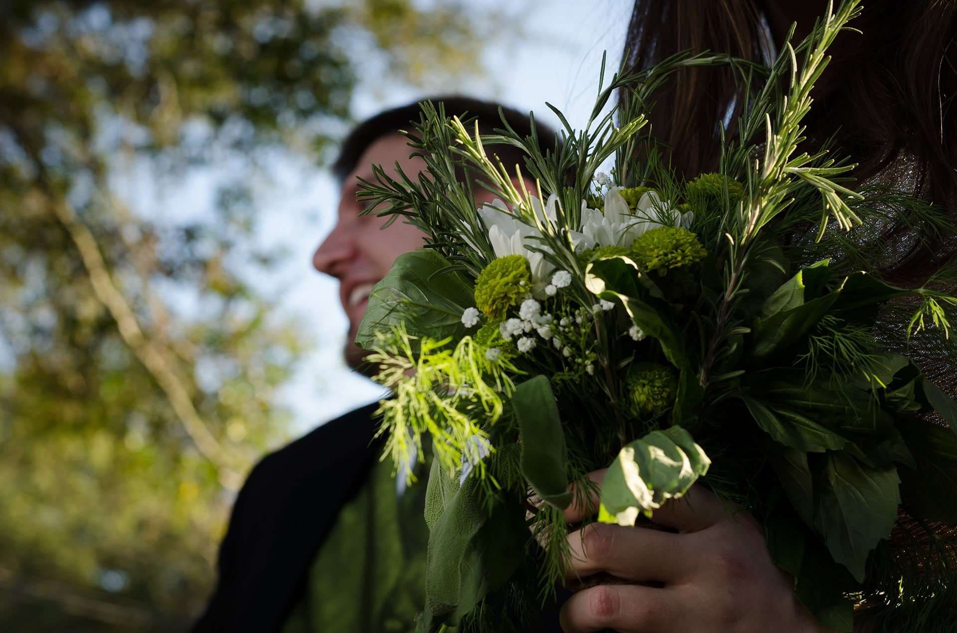 Smiling man in a dark jacket behind a green and white herb bouquet held in the foreground outdoors