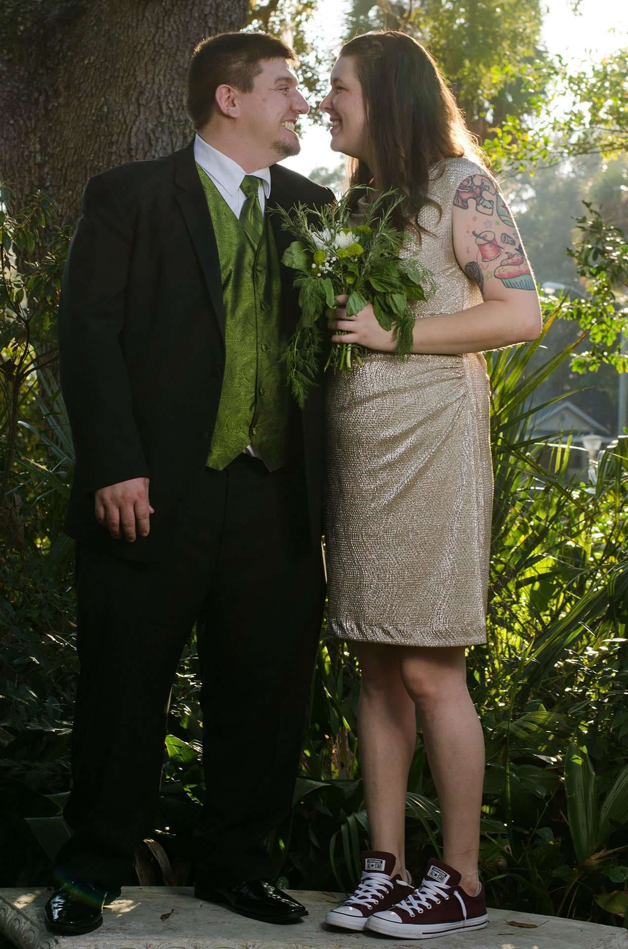 Couple smiling at each other outdoors, he in a black suit with green vest, she in a gold dress holding flowers