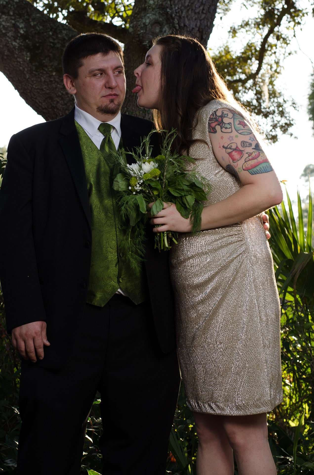 Woman with arm tattoos playfully sticks out her tongue beside a deadpan man in a green vest, holding a bouquet outdoors