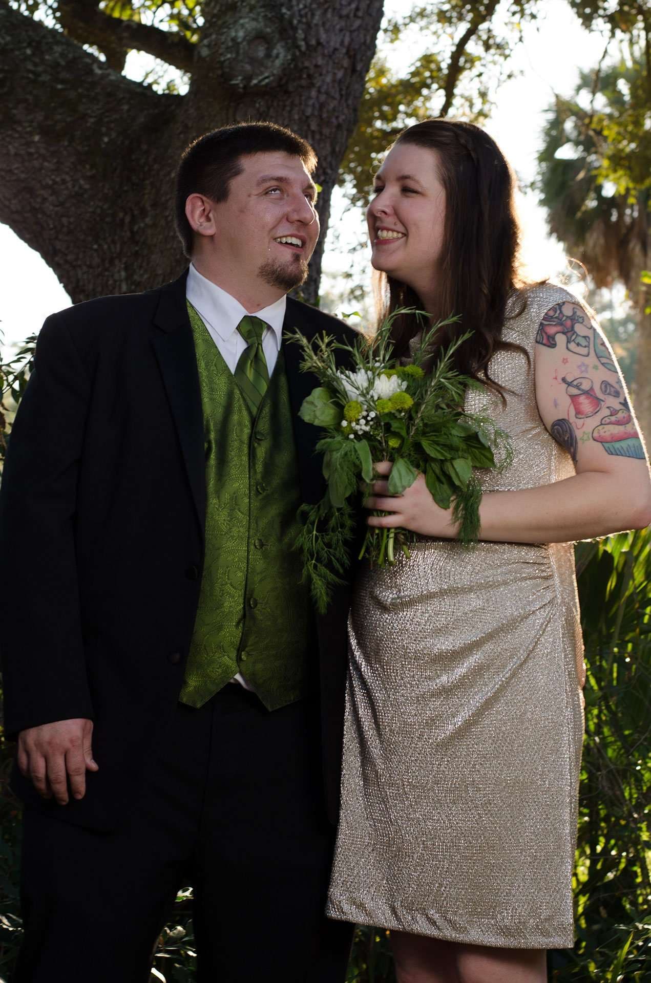 Smiling couple beneath an oak tree, man in black suit and green vest, woman in gold dress holding a white and green bouquet
