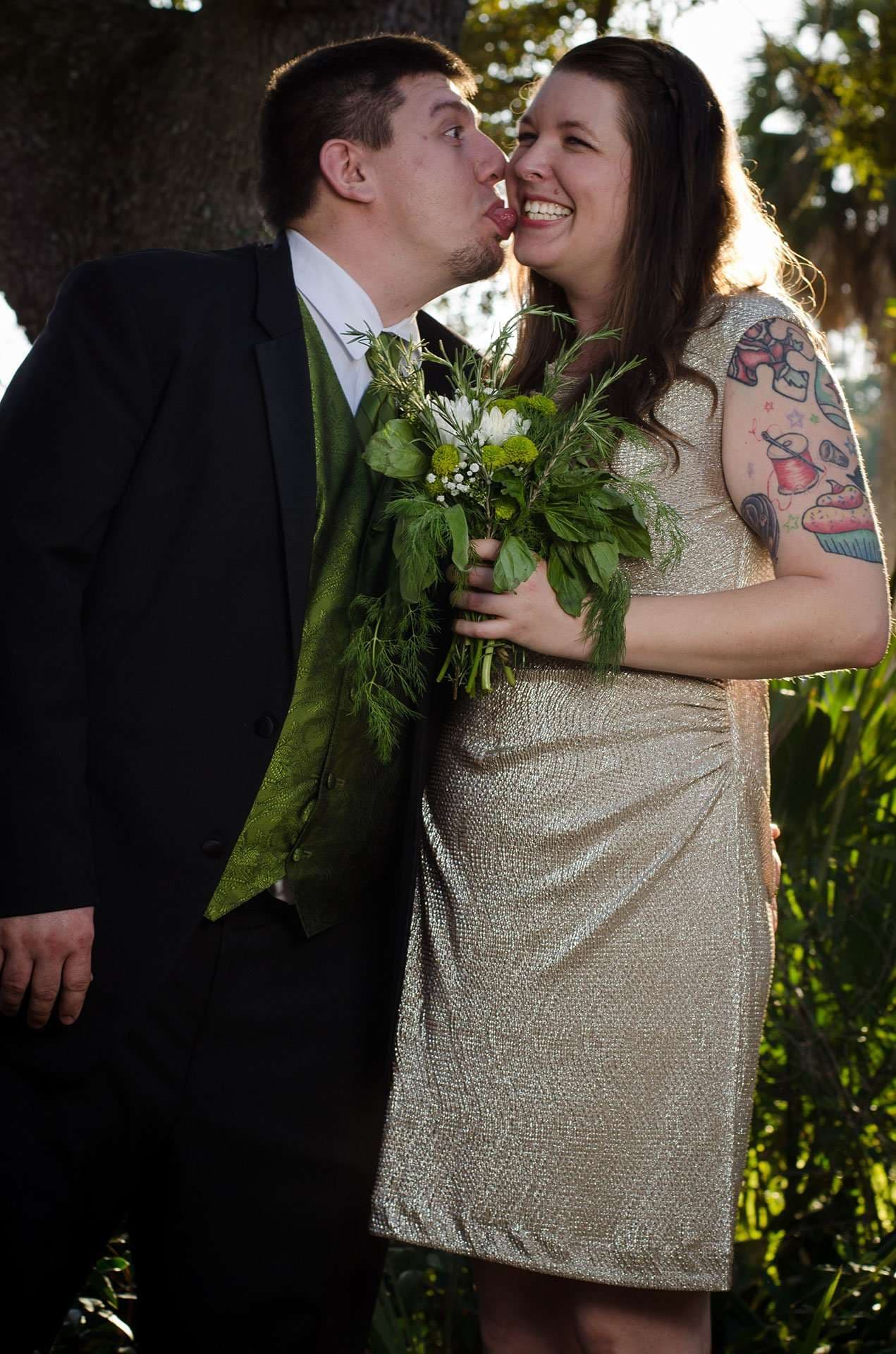Groom in dark suit and green vest playfully licks his laughing bride's cheek, she holds a green bouquet outdoors