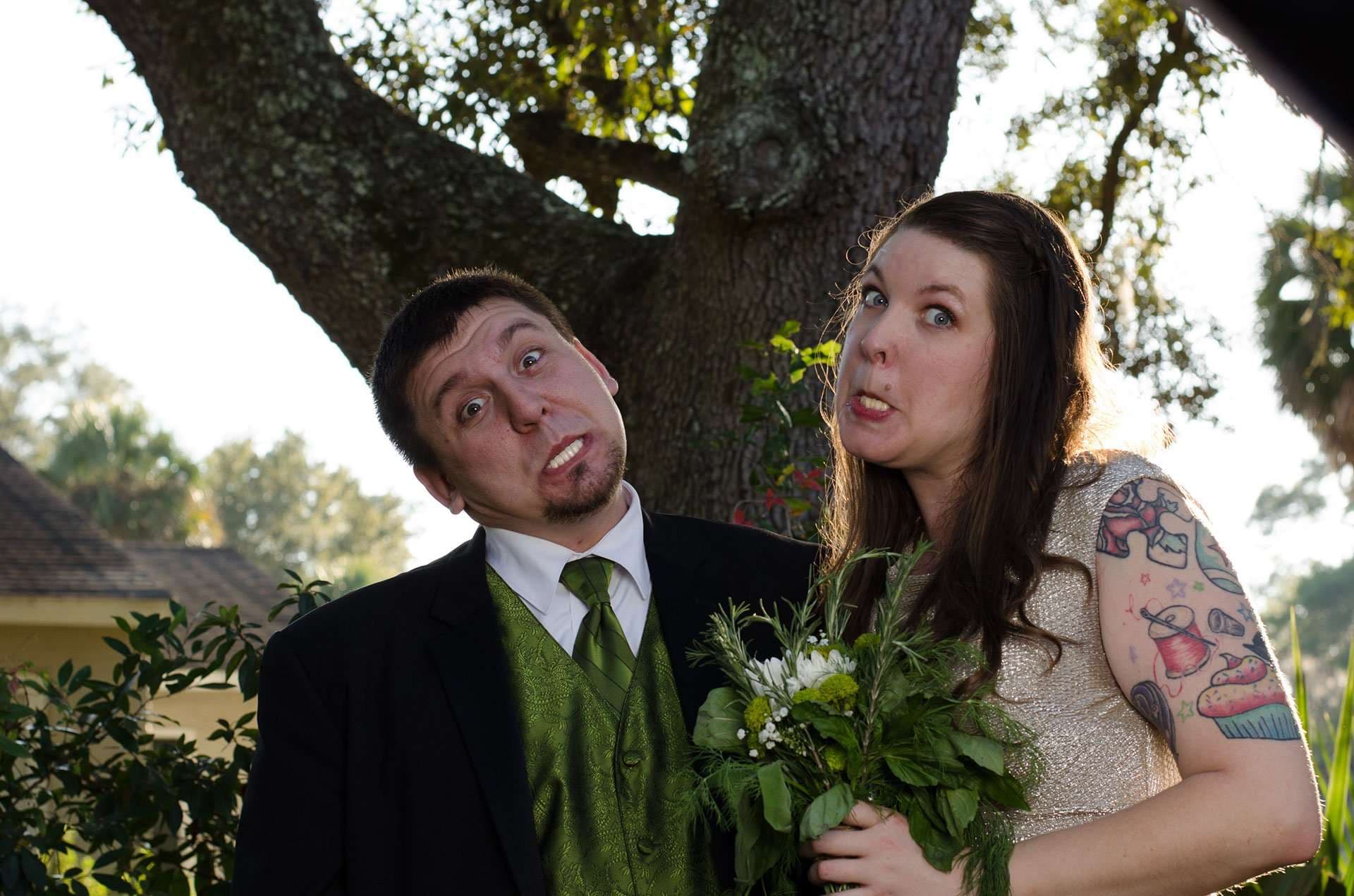 Couple making goofy faces under a large oak tree, he in a green vest and tie, she holding a green bouquet