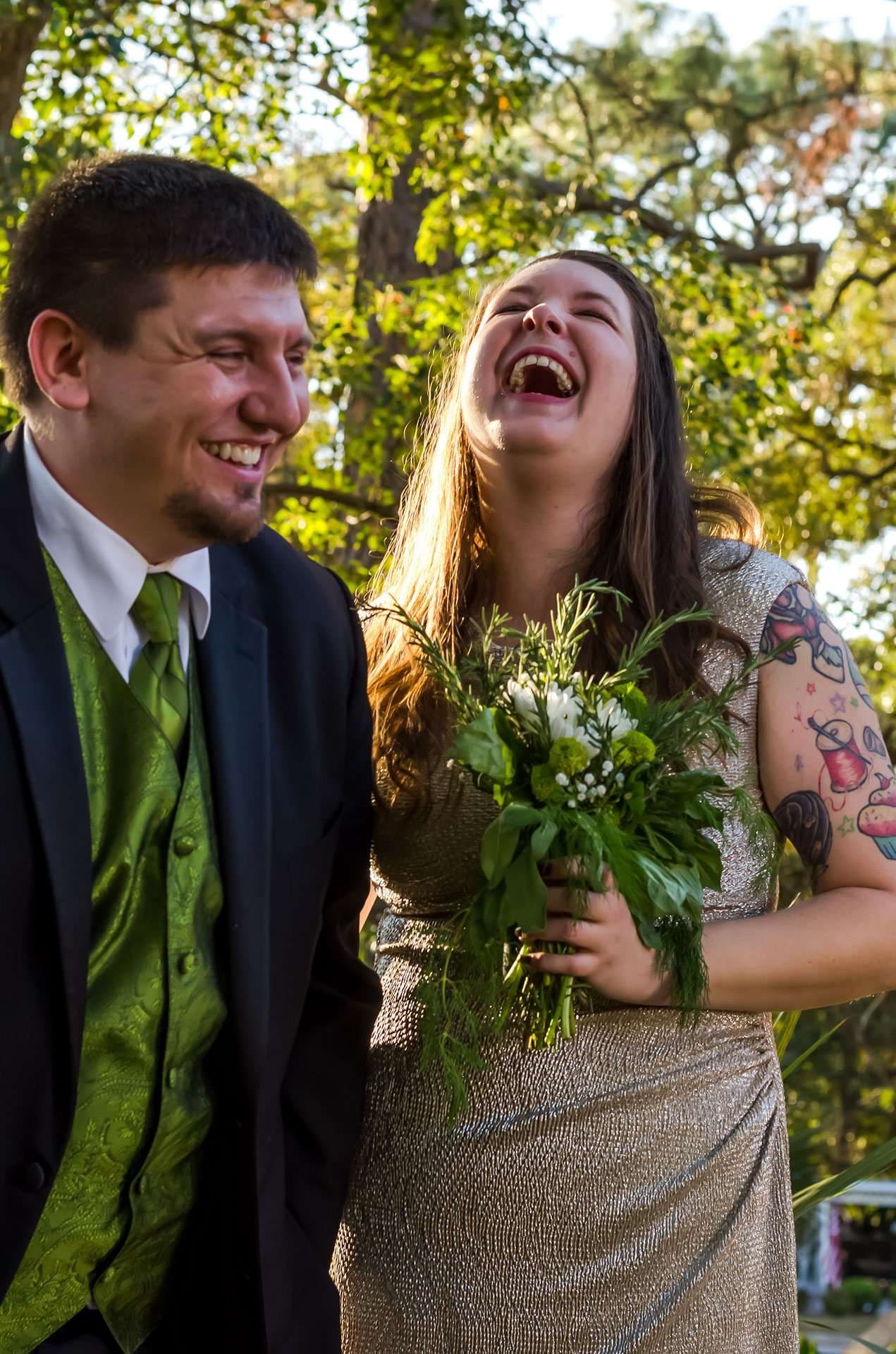 Couple laughing outdoors under trees, the groom in a green paisley vest and the woman with tattoos holding a green bouquet
