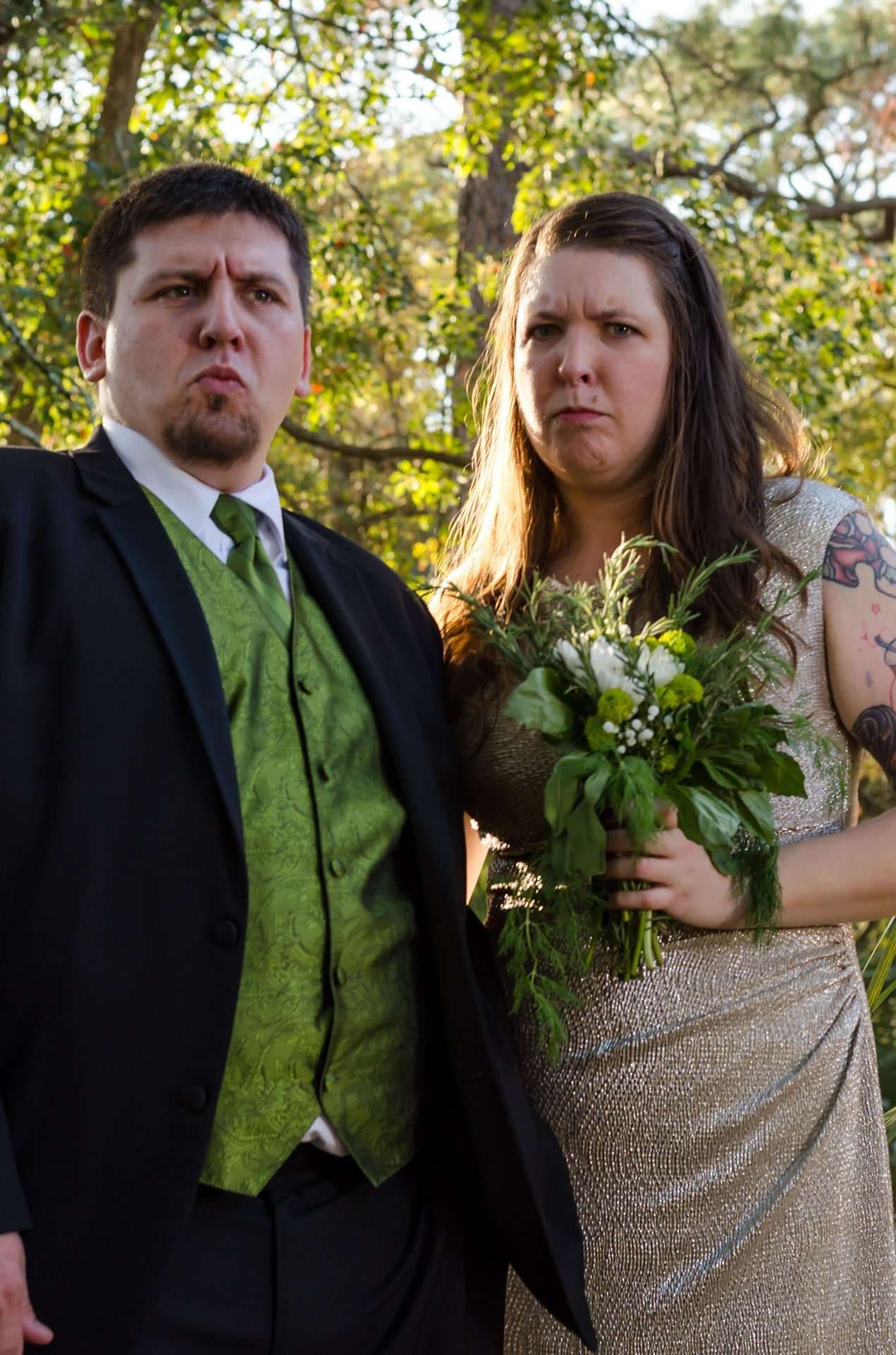Couple in formal wedding attire making comically stern faces outdoors under trees, she holds a green and white bouquet