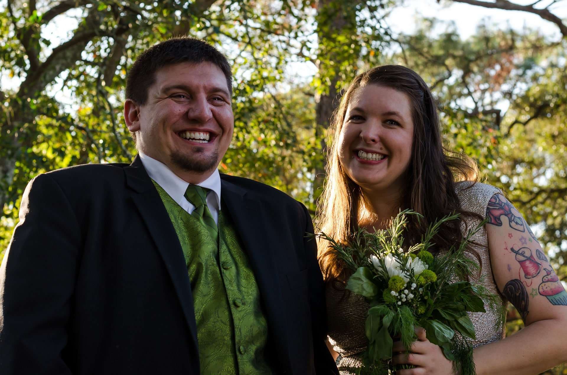 Smiling couple outdoors under trees, man in black jacket and green vest, woman with tattooed arm holding a bouquet