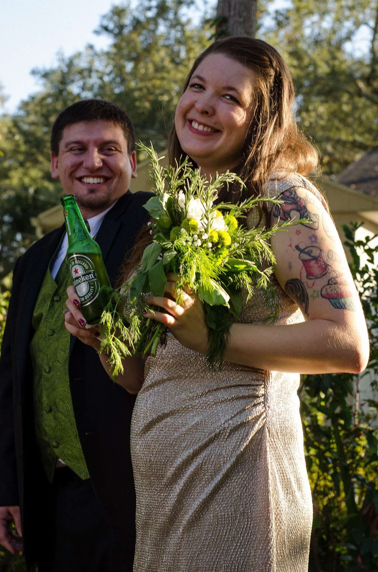 Smiling couple outdoors, she holds a green and white bouquet, he holds a Heineken bottle in a green vest