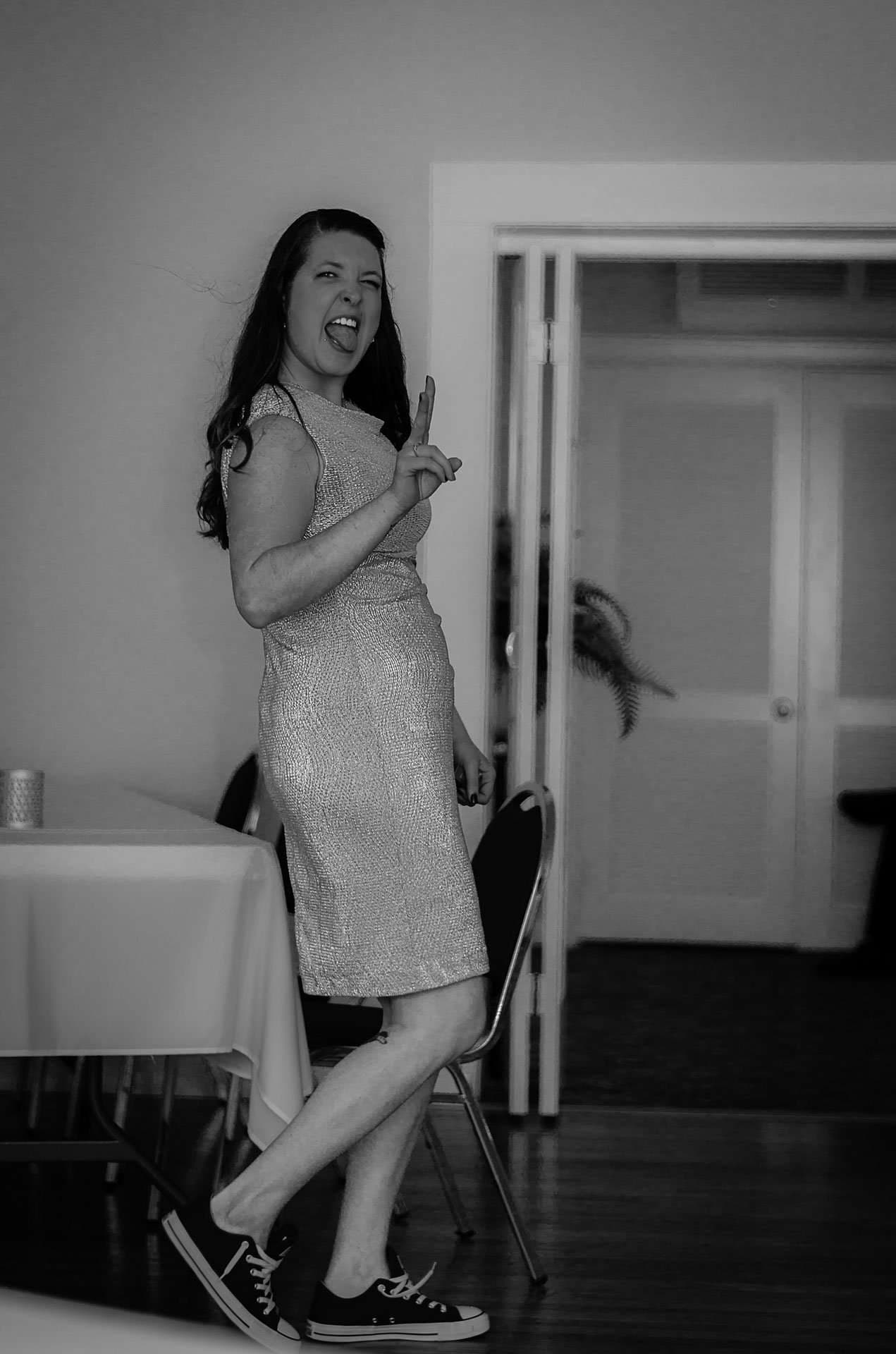 Black-and-white shot of a woman in a shimmery dress and sneakers sticking out her tongue and winking indoors