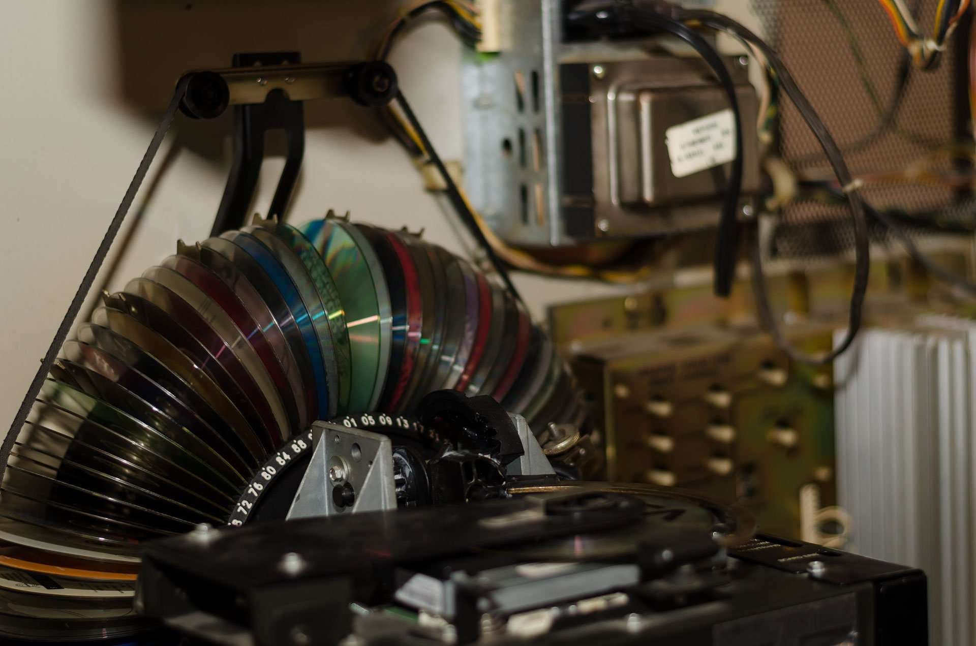 Curved stack of compact discs fanned along a track inside a jukebox mechanism, with wires and a transformer behind
