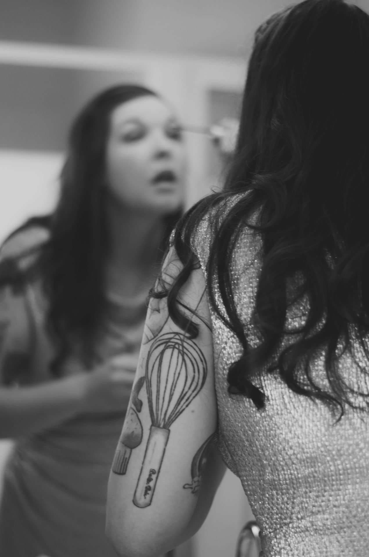Black-and-white shot of a woman applying mascara in a mirror, her forearm tattooed with a whisk and kitchen utensils