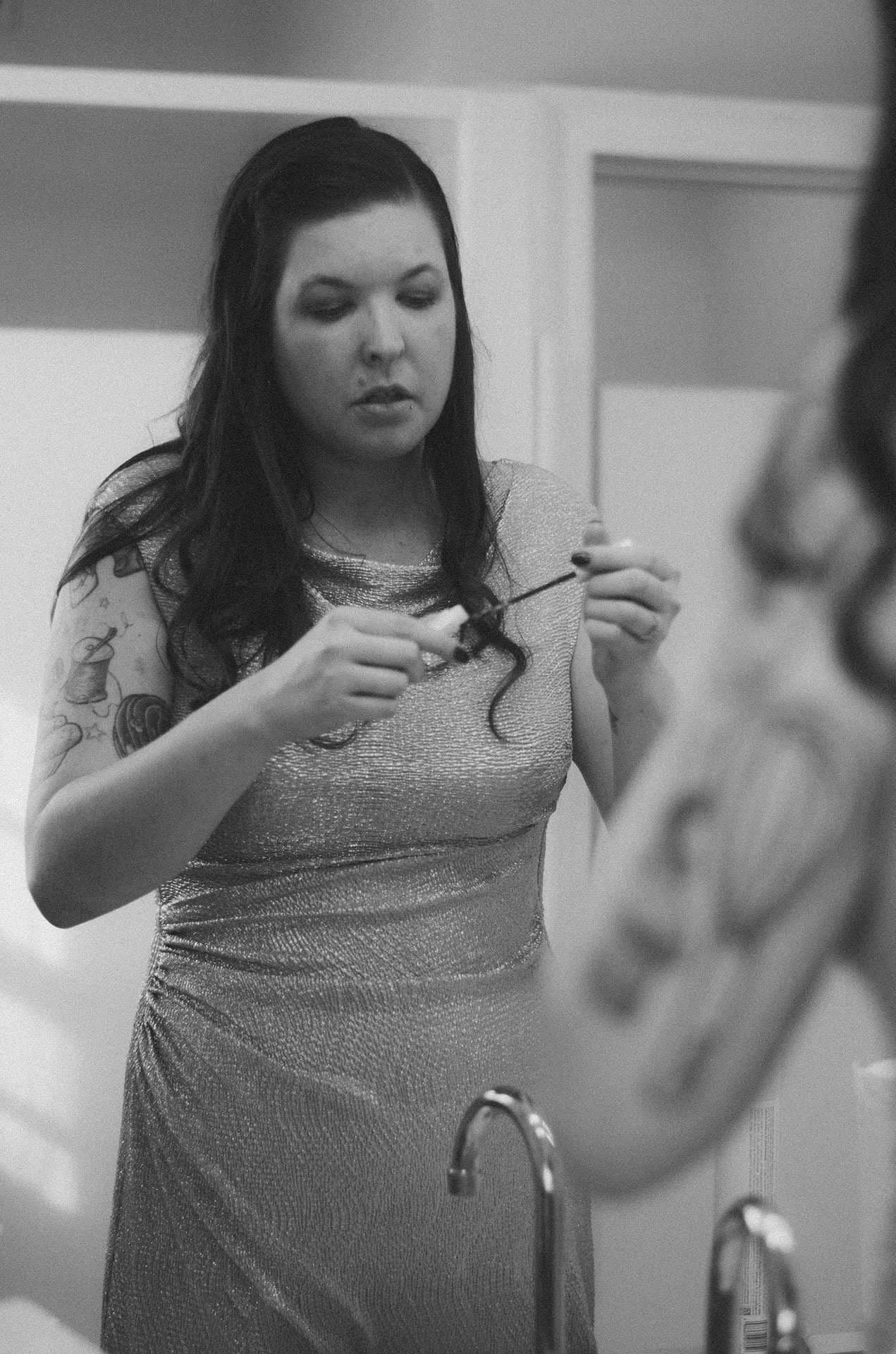 Black-and-white photo of a woman in a shimmery dress applying mascara while looking into a mirror, tattoo on her arm