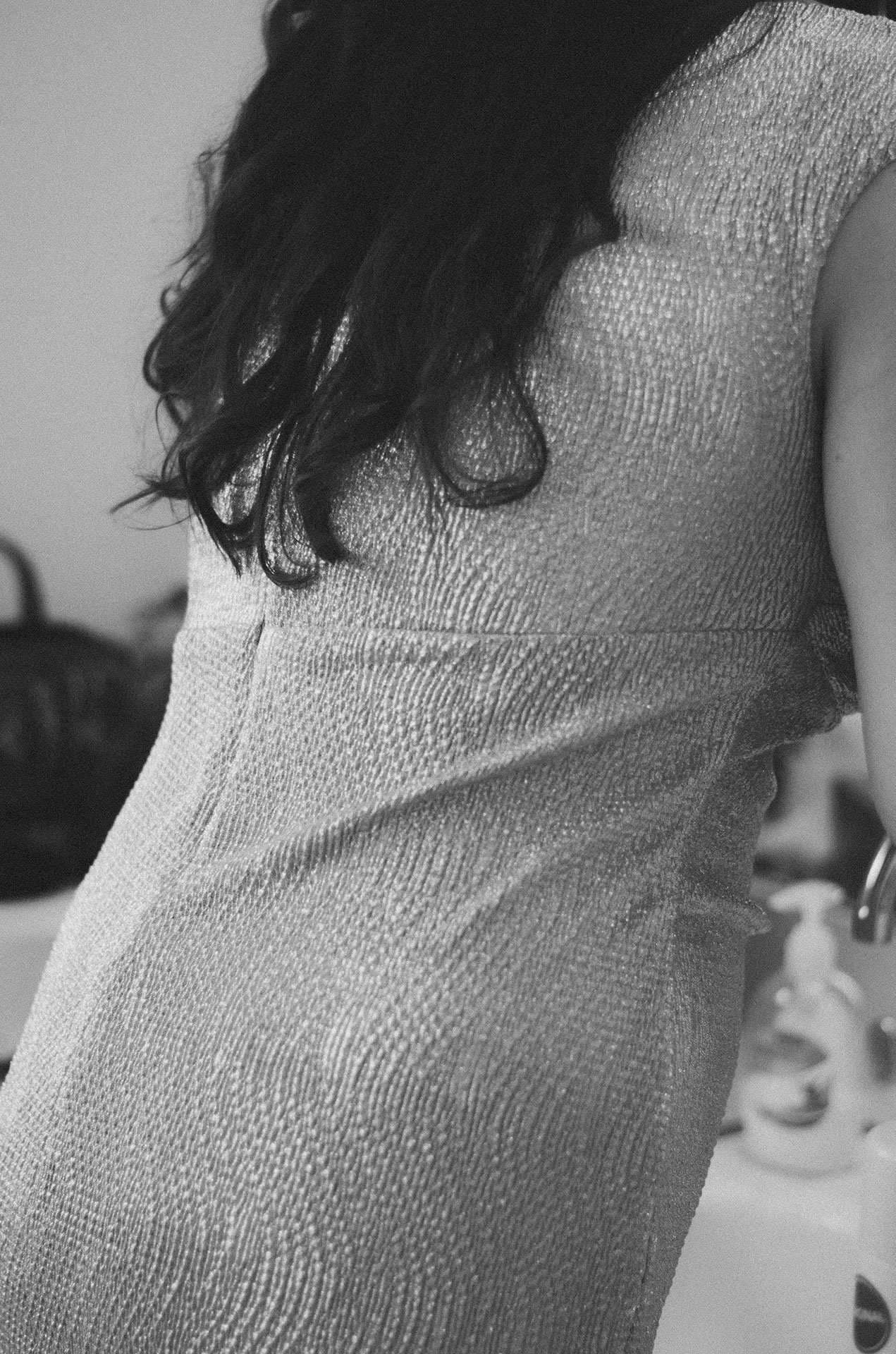 Black-and-white close-up of a woman's back in a shimmery textured dress, dark wavy hair falling over her shoulder
