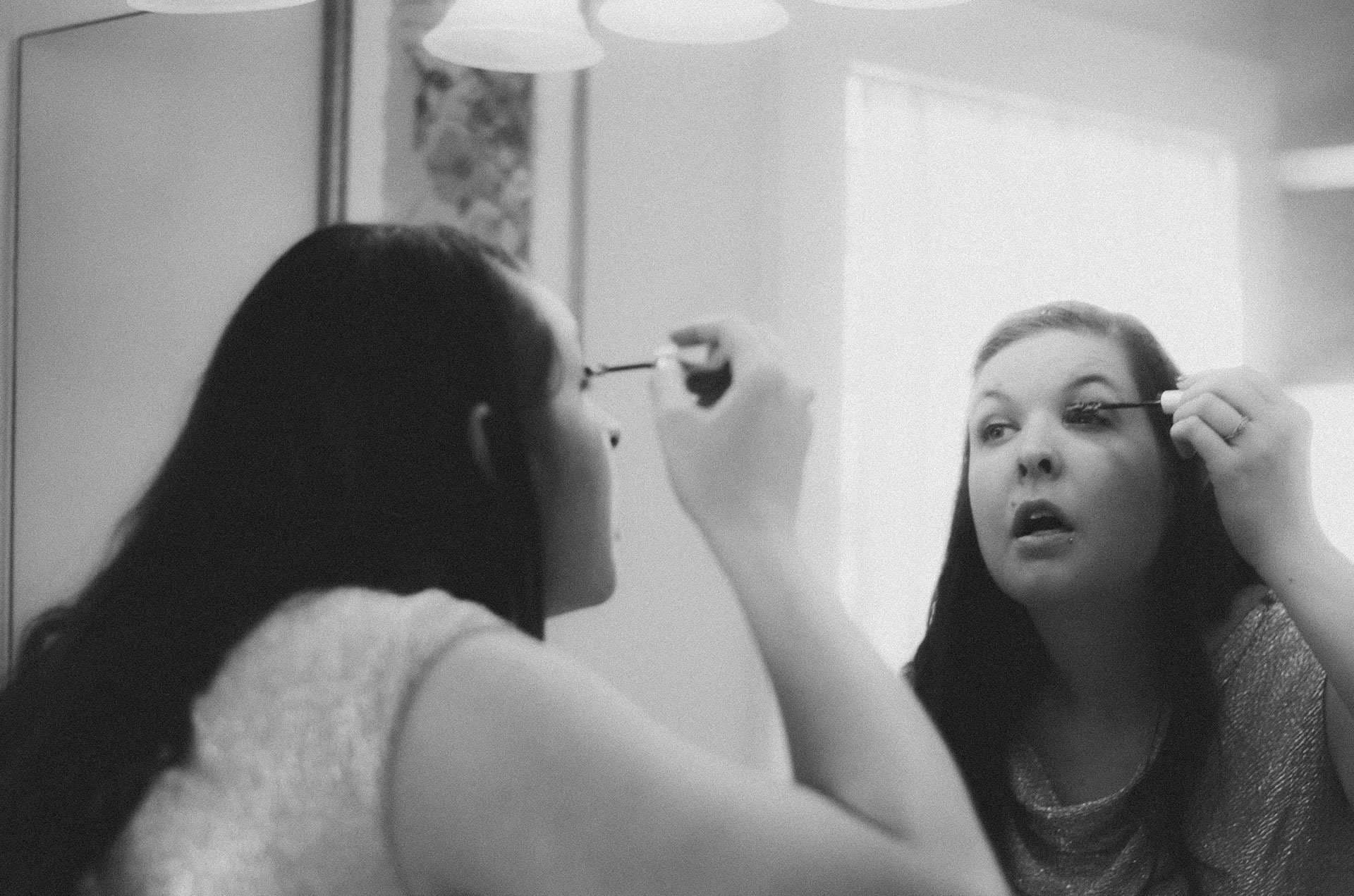 Black-and-white shot of a woman applying mascara while looking into a mirror, her reflection facing us