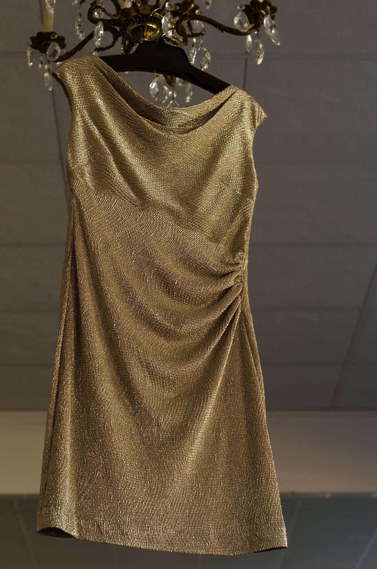 Gold metallic sleeveless cocktail dress with gathered side ruching hanging on a dark hanger beneath a crystal chandelier