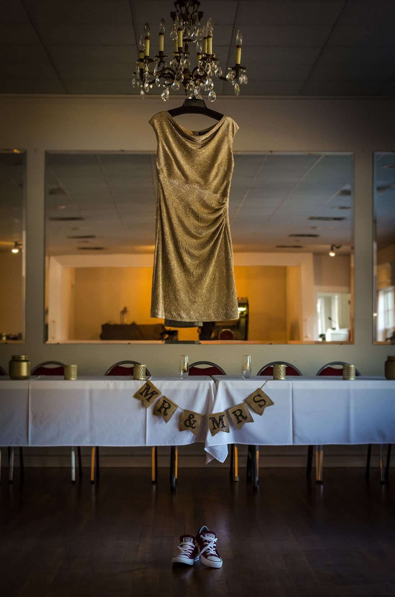 Gold sequin dress hanging from a chandelier above a Mr. & Mrs. banner, head table and red sneakers on the floor
