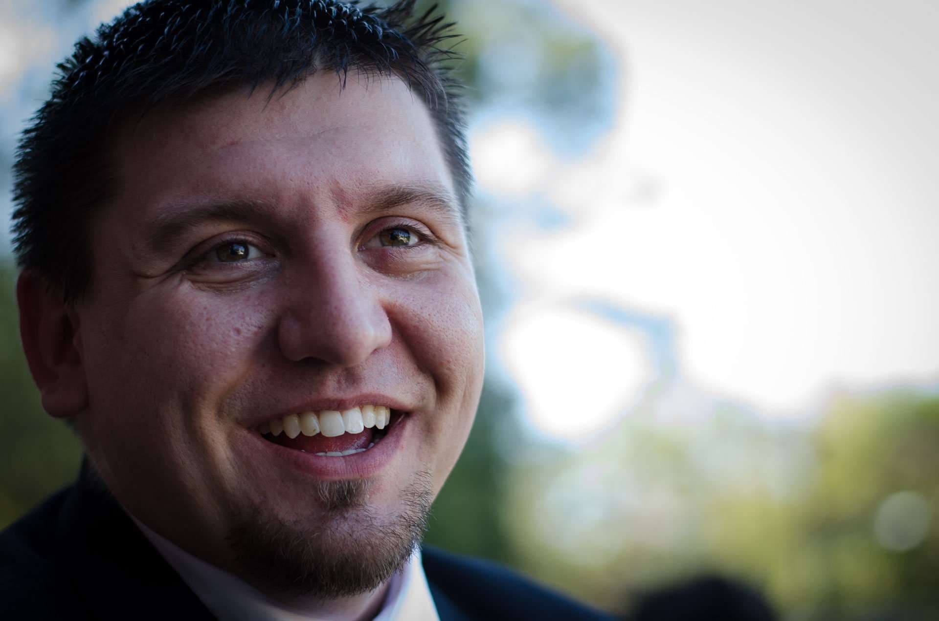 Smiling man with short dark hair and a goatee in a dark suit, outdoors with blurred trees behind him