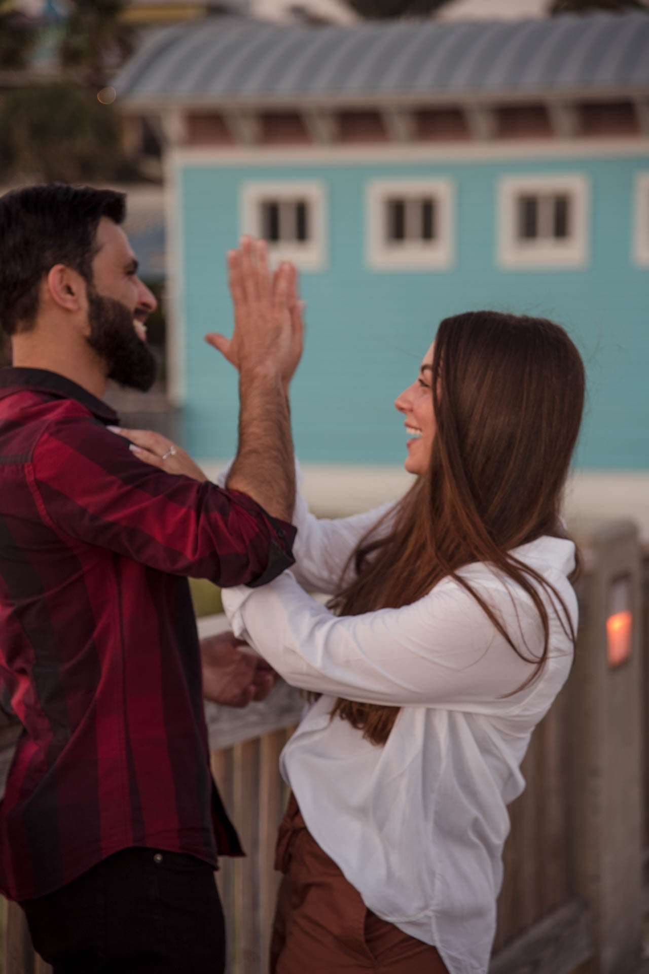Smiling couple high-fiving on a wooden boardwalk, woman in a white blouse, man in a plaid shirt, blue building behind