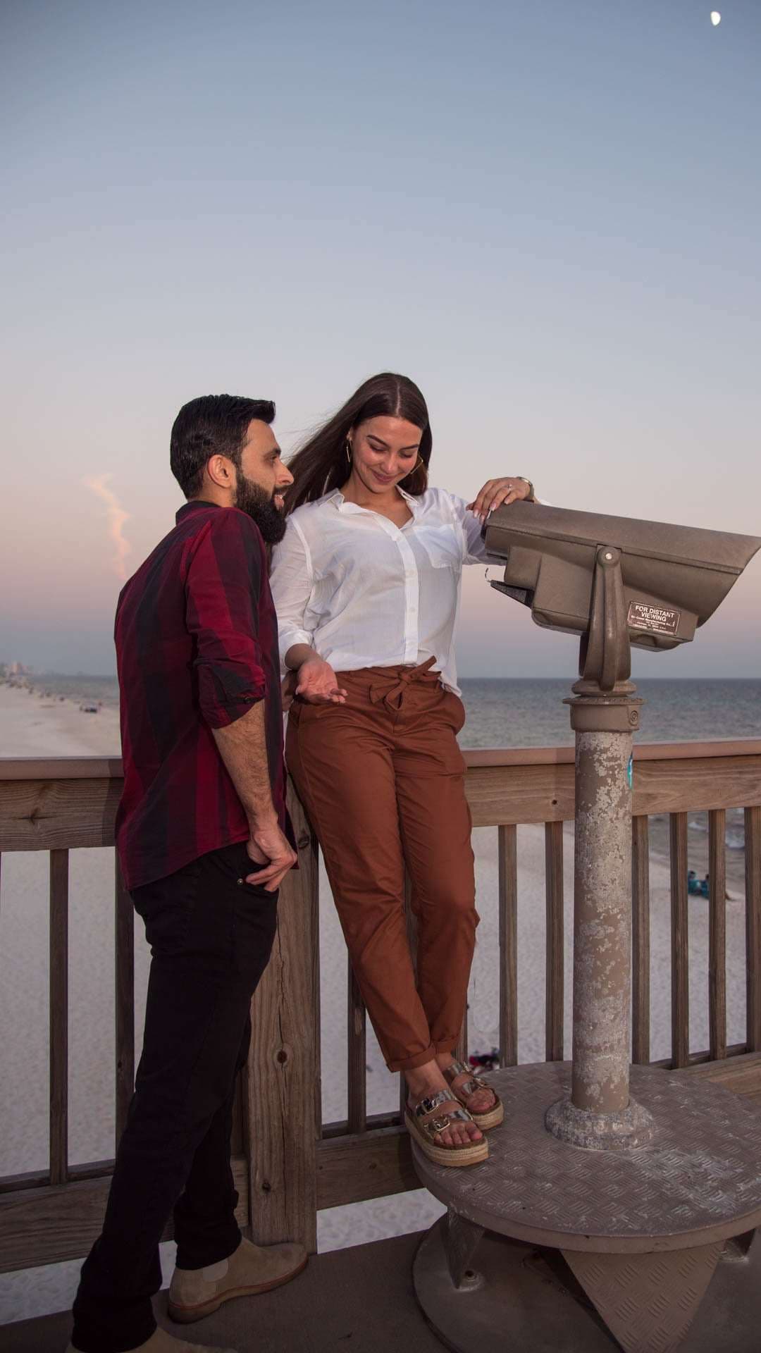 Smiling woman leaning on a pier railing beside a bearded man, with a coin-operated viewer and beach at dusk