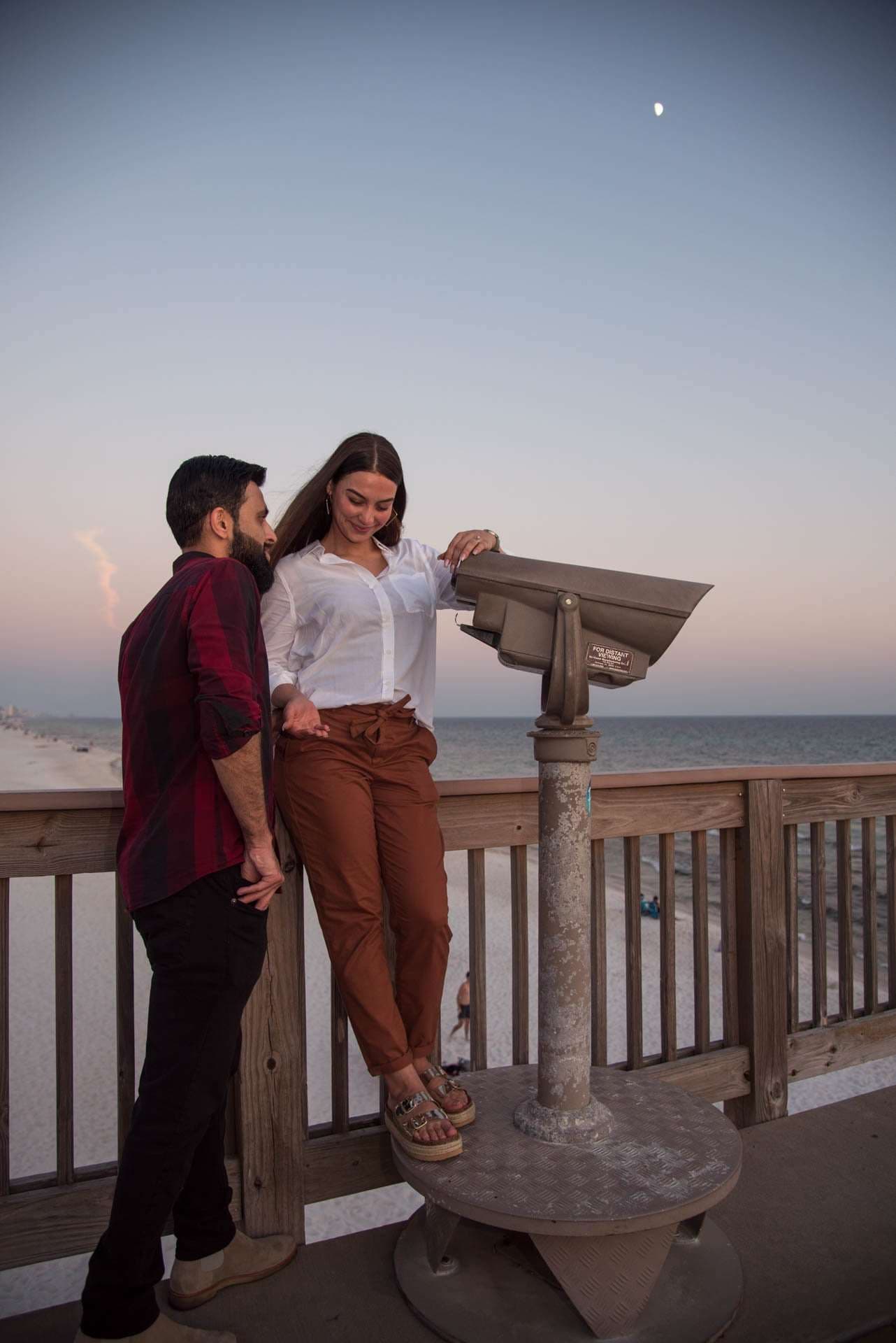 Smiling couple leaning together beside a coin-operated viewer on the Panama City Beach City Pier at dusk, moon above