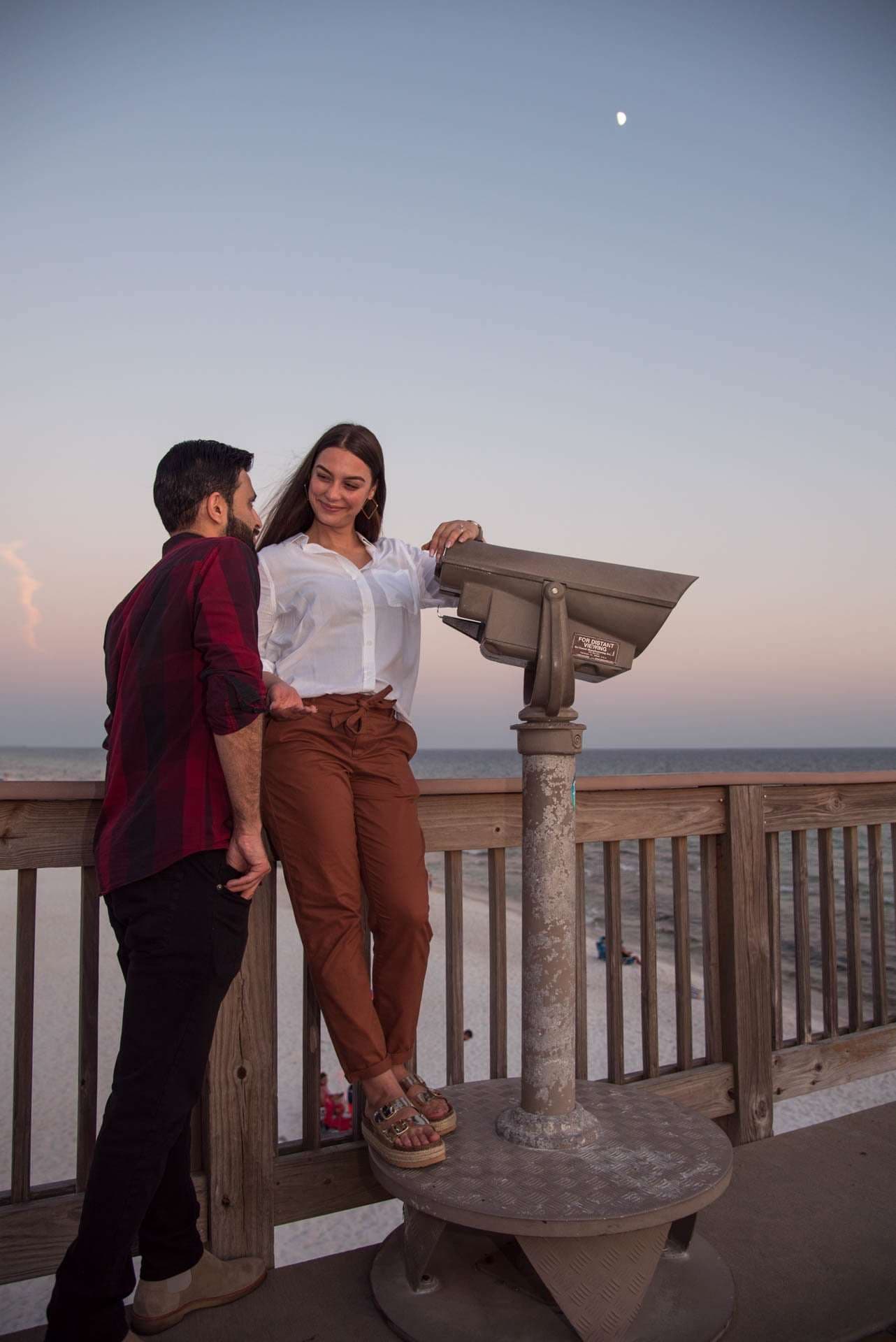 Smiling woman standing on a viewer stand leans toward a man beside her on the Panama City Beach City Pier at dusk