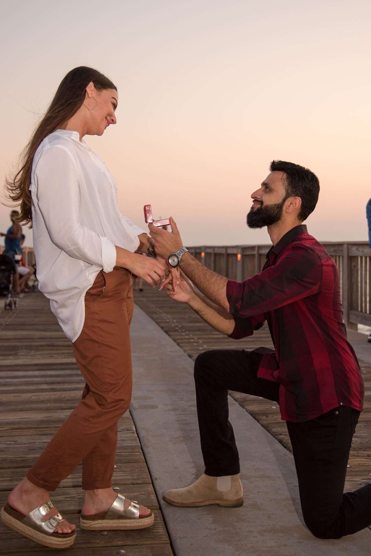 Man kneeling on a wooden pier holding out a ring box to a smiling woman at Panama City Beach City Pier at sunset