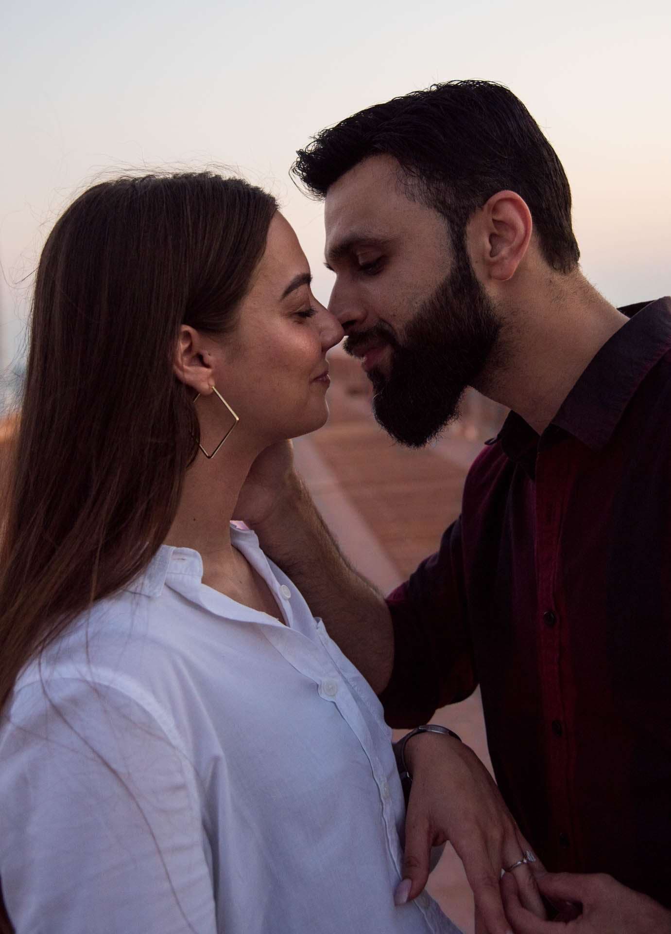Couple touching noses before a kiss on the pier at dusk, woman in white shirt, bearded man in maroon