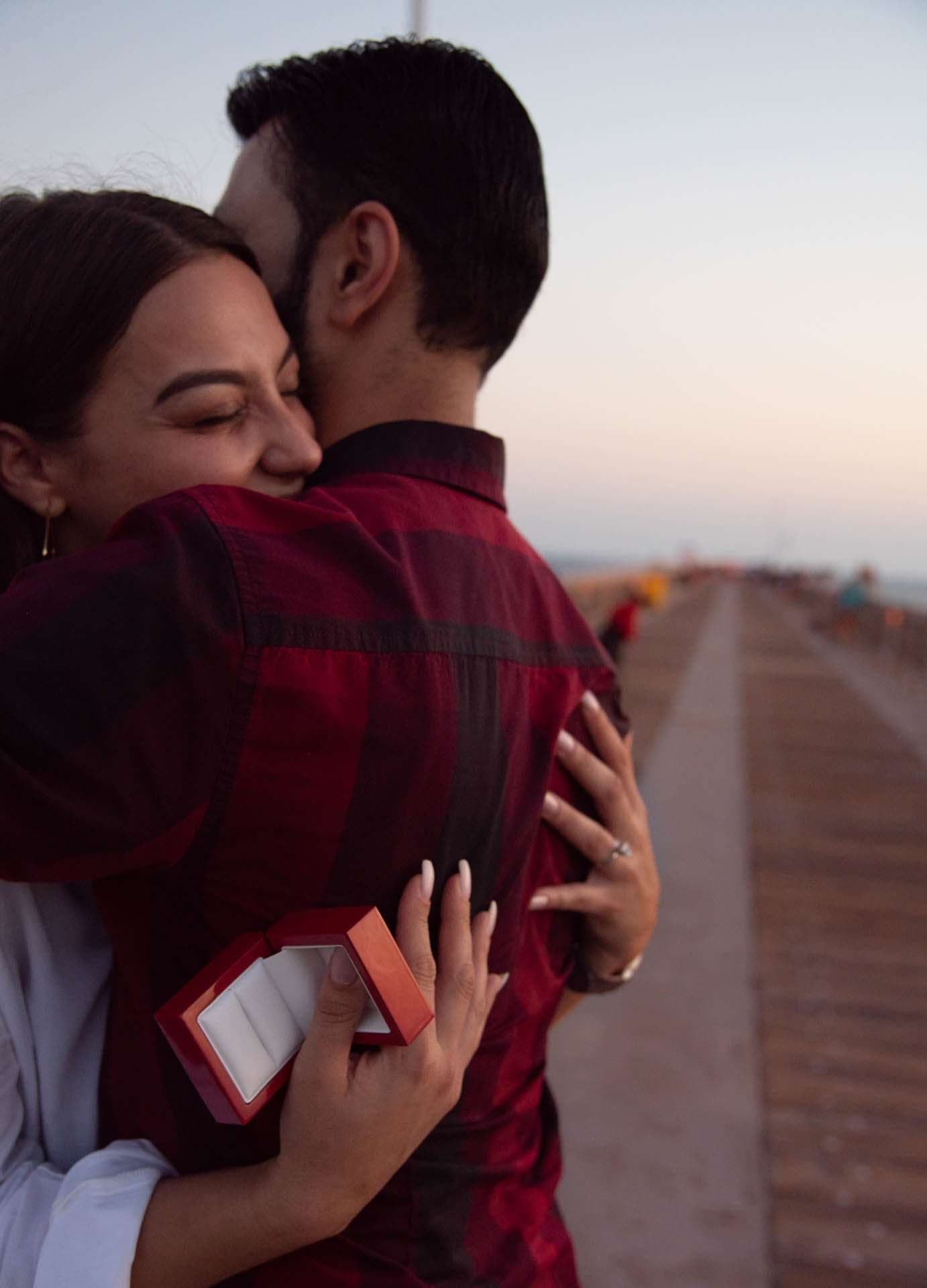 Woman hugging her new fiancé and holding an open red ring box on the Panama City Beach City Pier at dusk